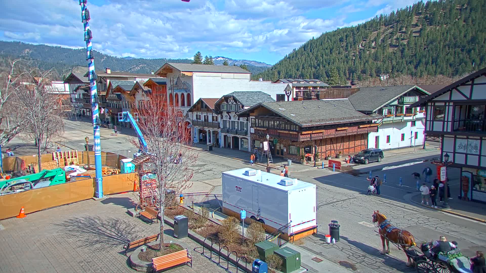 A mostly sunny day shows a town square with Bavarian-style buildings, a horse-drawn carriage, and construction in progress, all nestled in a valley with forested hillsides.