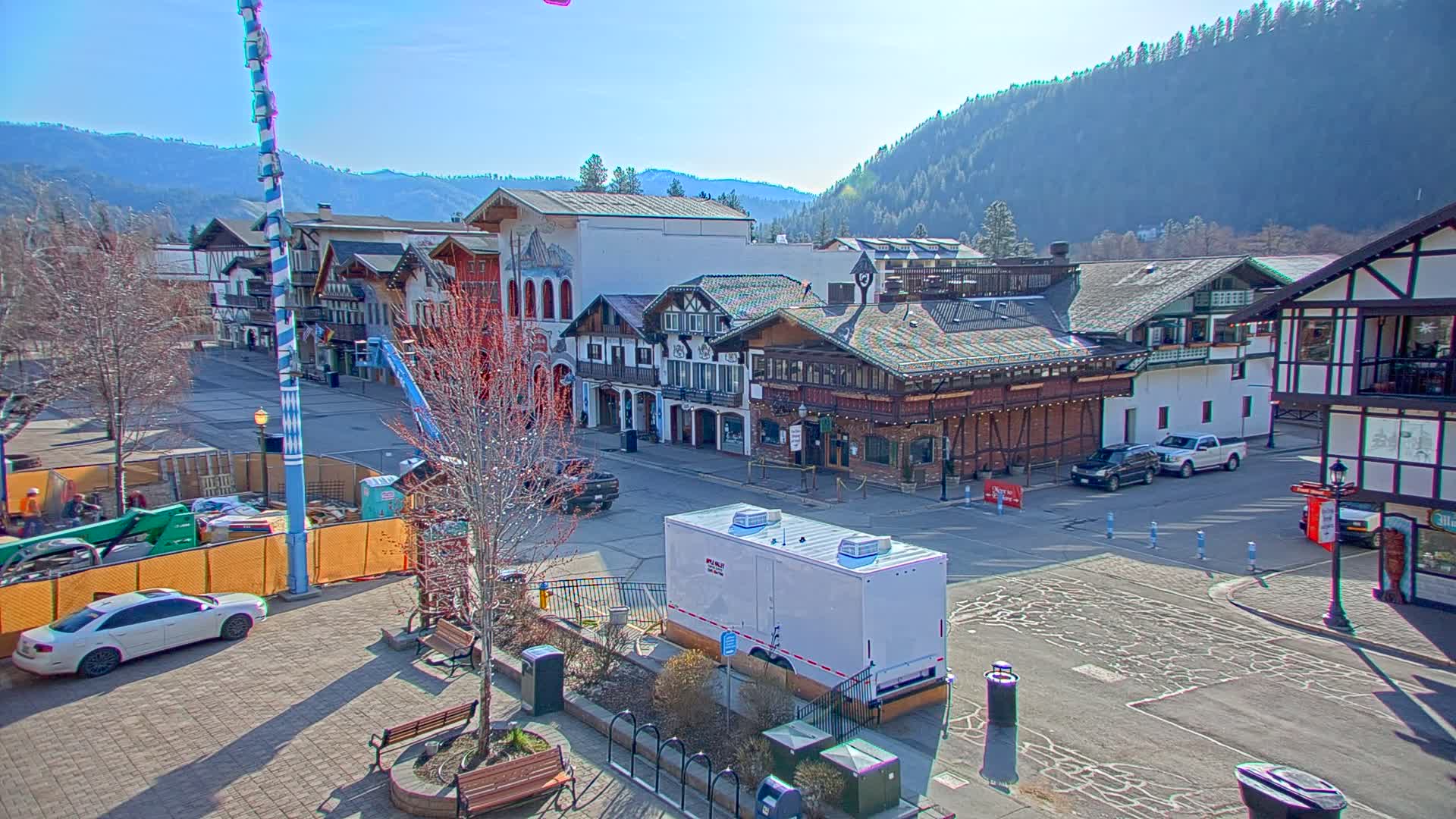 A sunny day reveals a Bavarian-style town square with several buildings, some vehicles parked, and construction activity in progress.