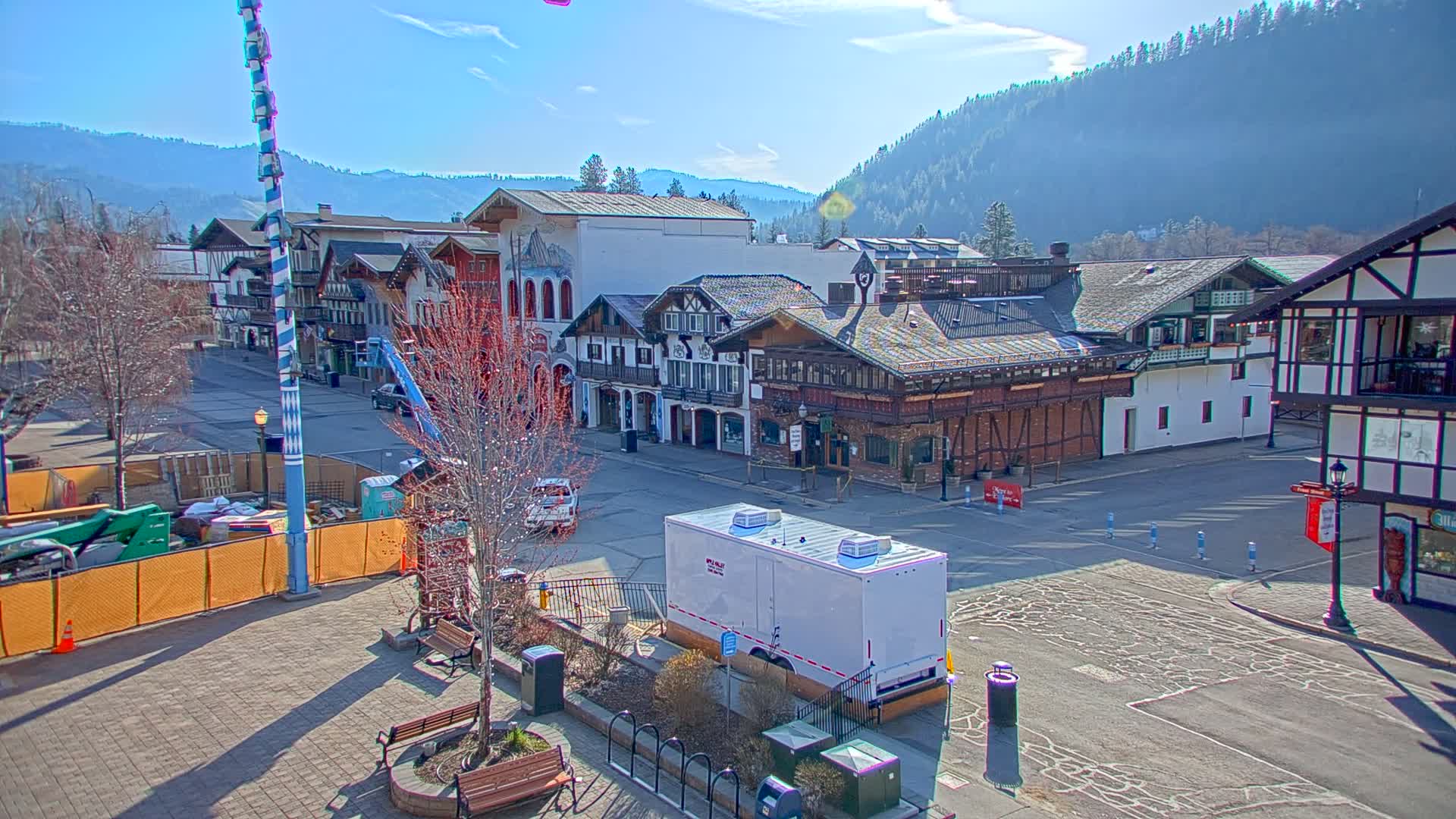 A sunny day reveals a town square with several Bavarian-style buildings, construction barriers, and a large white trailer, all situated against a backdrop of a mountain range.