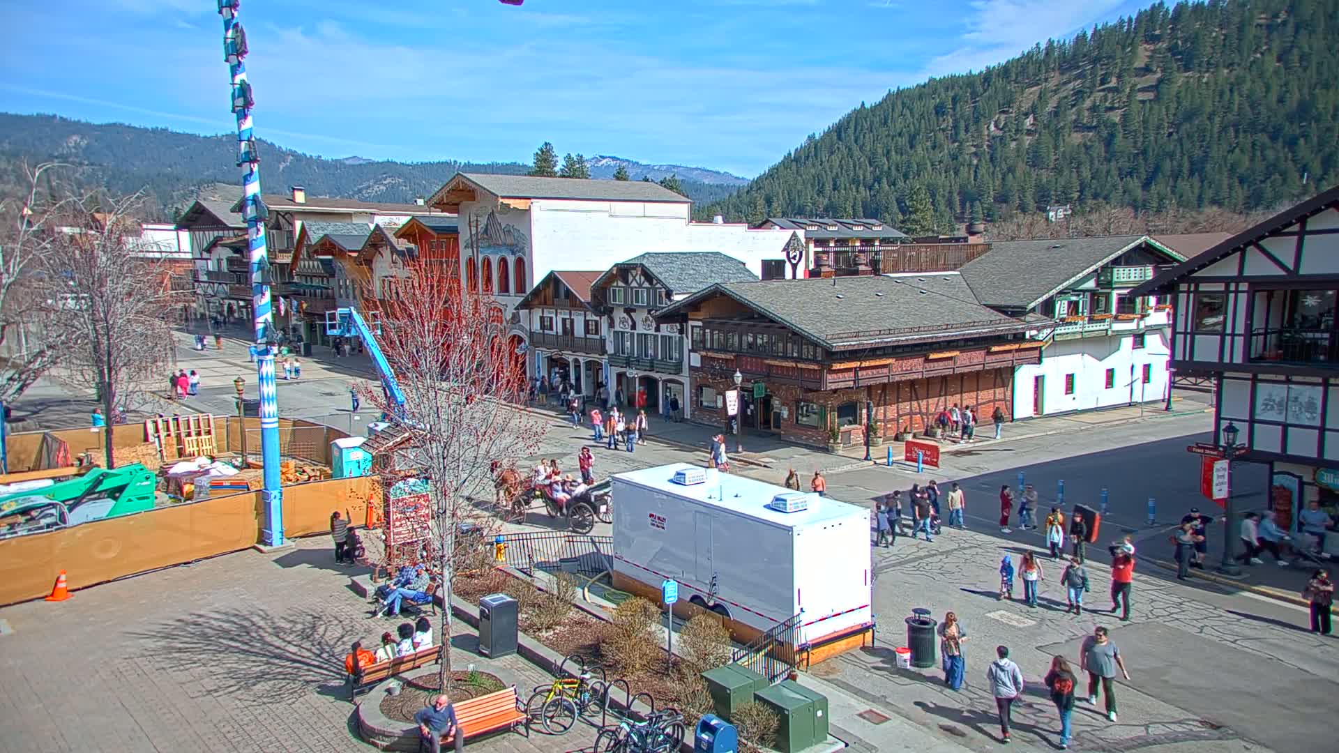 A sunny day in a Bavarian-style town square with several people walking around, buildings lining the street, and construction occurring in a fenced-off area.