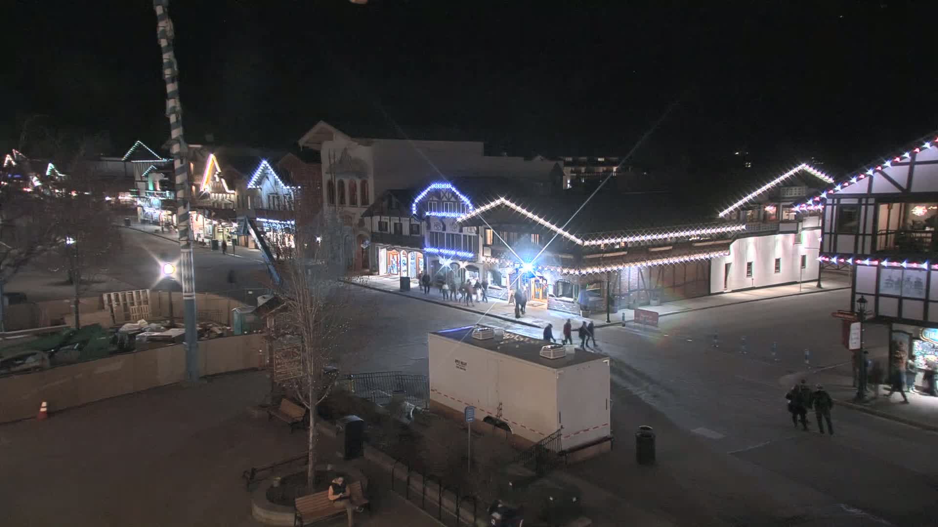 A nighttime view of a town square with several buildings decorated with string lights, some people walking around, and a construction area visible in the foreground.