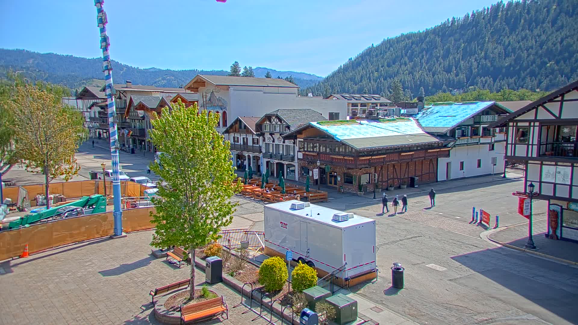 A town square with Bavarian-style buildings, outdoor seating, and a tall maypole under a clear, sunny sky, backed by a mountain range covered in dark green trees.