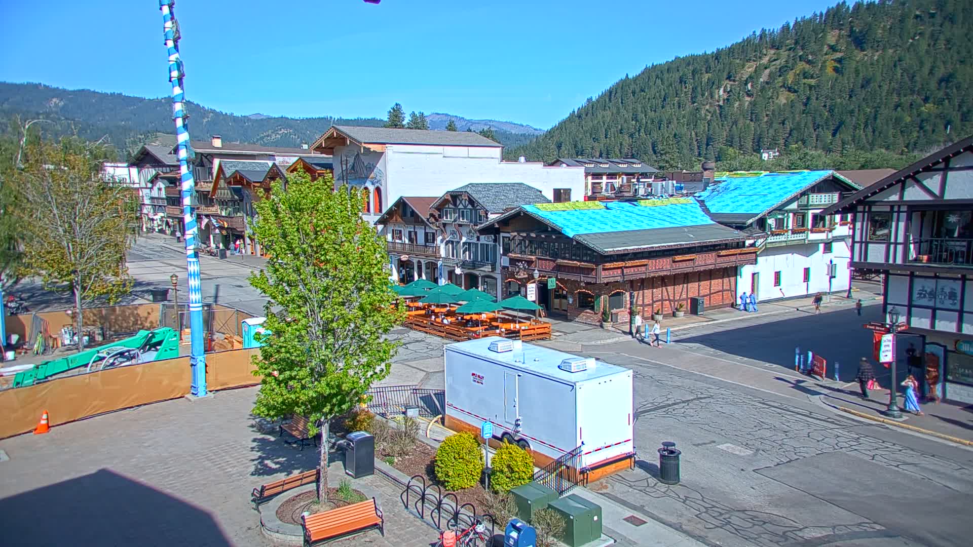 A town square with Bavarian-style buildings, outdoor seating, and a partially-constructed structure, all under a clear, sunny sky and surrounded by a mountain range.