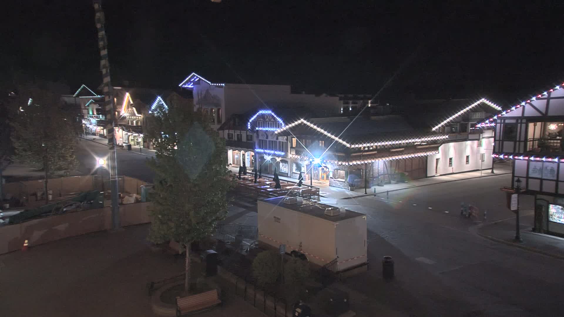 A nighttime view of a street lined with festively lit buildings, a portable restroom, and some trees.
