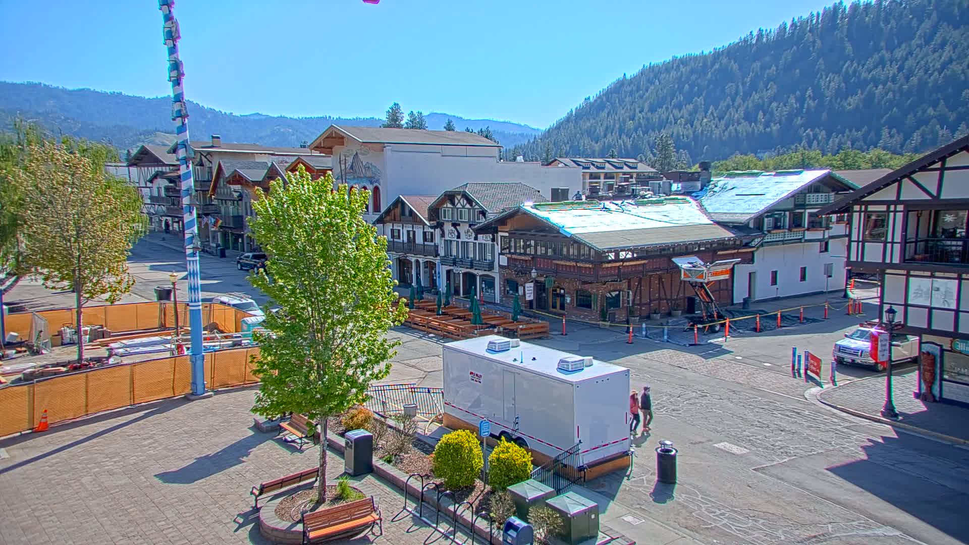 A town square with Bavarian-style buildings under a sunny sky, featuring construction in progress and a portable restroom.
