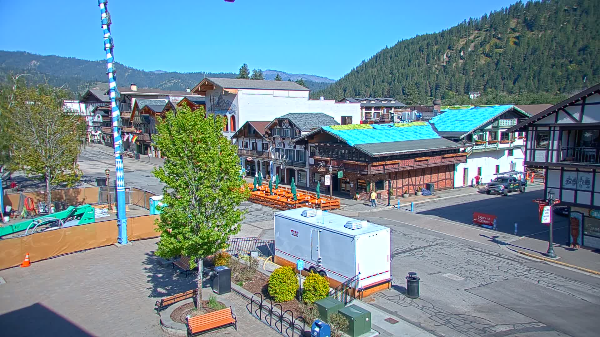 A town square with European-style buildings, outdoor seating, and a tall maypole is situated against a backdrop of a mountain range under a clear, sunny sky.