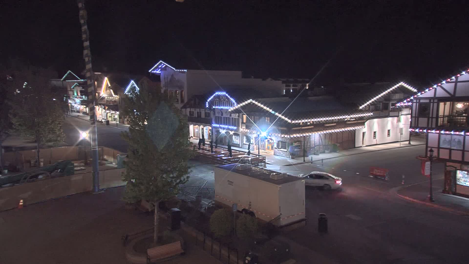 A nighttime, street-level view of a row of festively lit buildings, a vehicle driving on the street, and a trailer parked nearby.