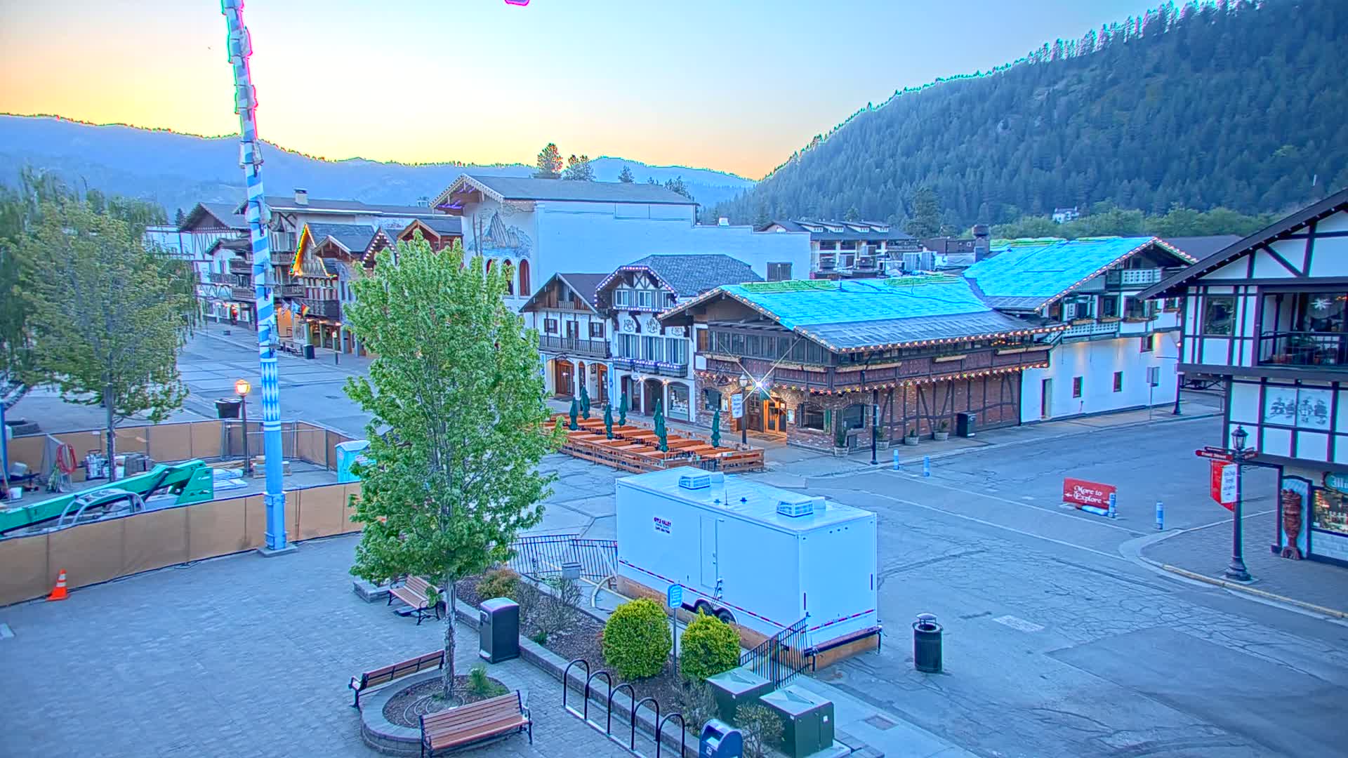A mostly empty town square with Bavarian-style buildings, outdoor seating, and a tall blue and white pole, under a clear sunrise sky and mountains in the background.