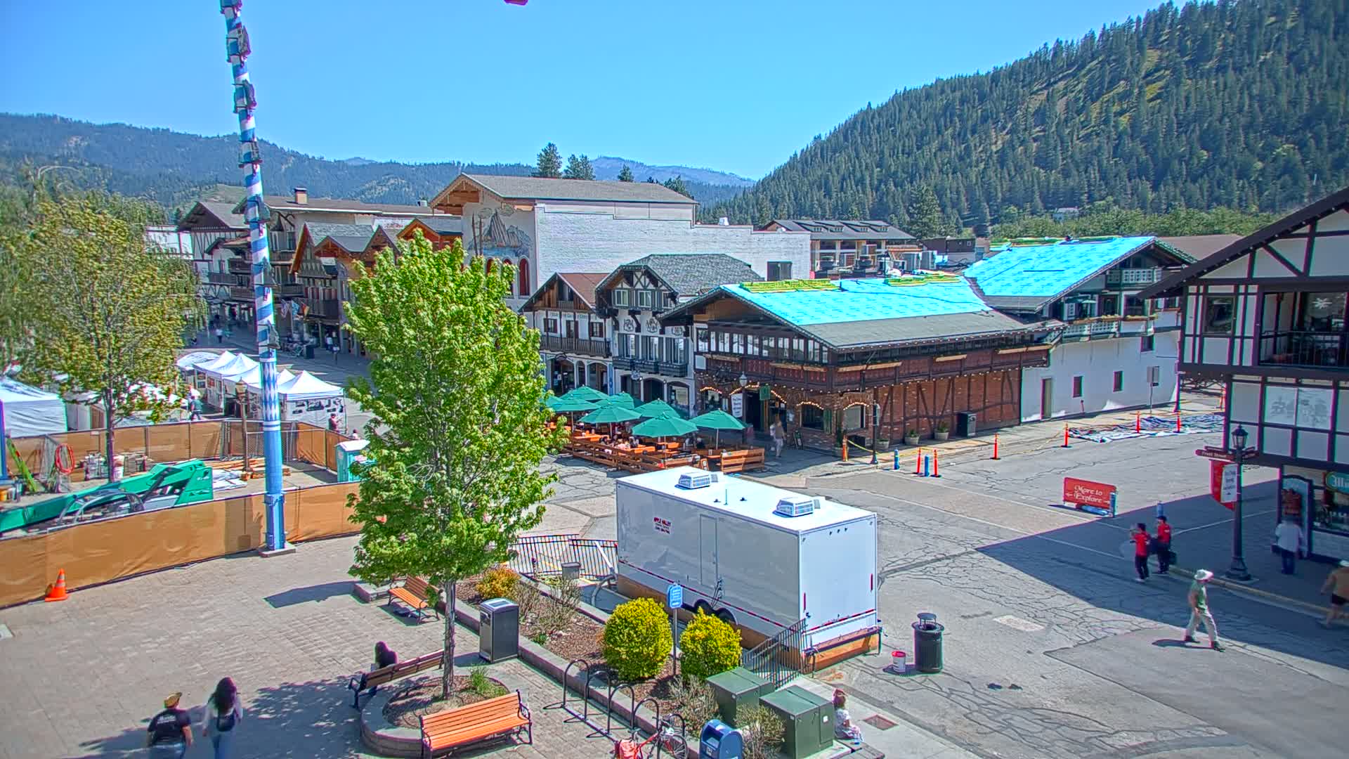 A town square with European-style buildings, outdoor seating, and a Maypole, under a sunny sky and surrounded by forested hills.