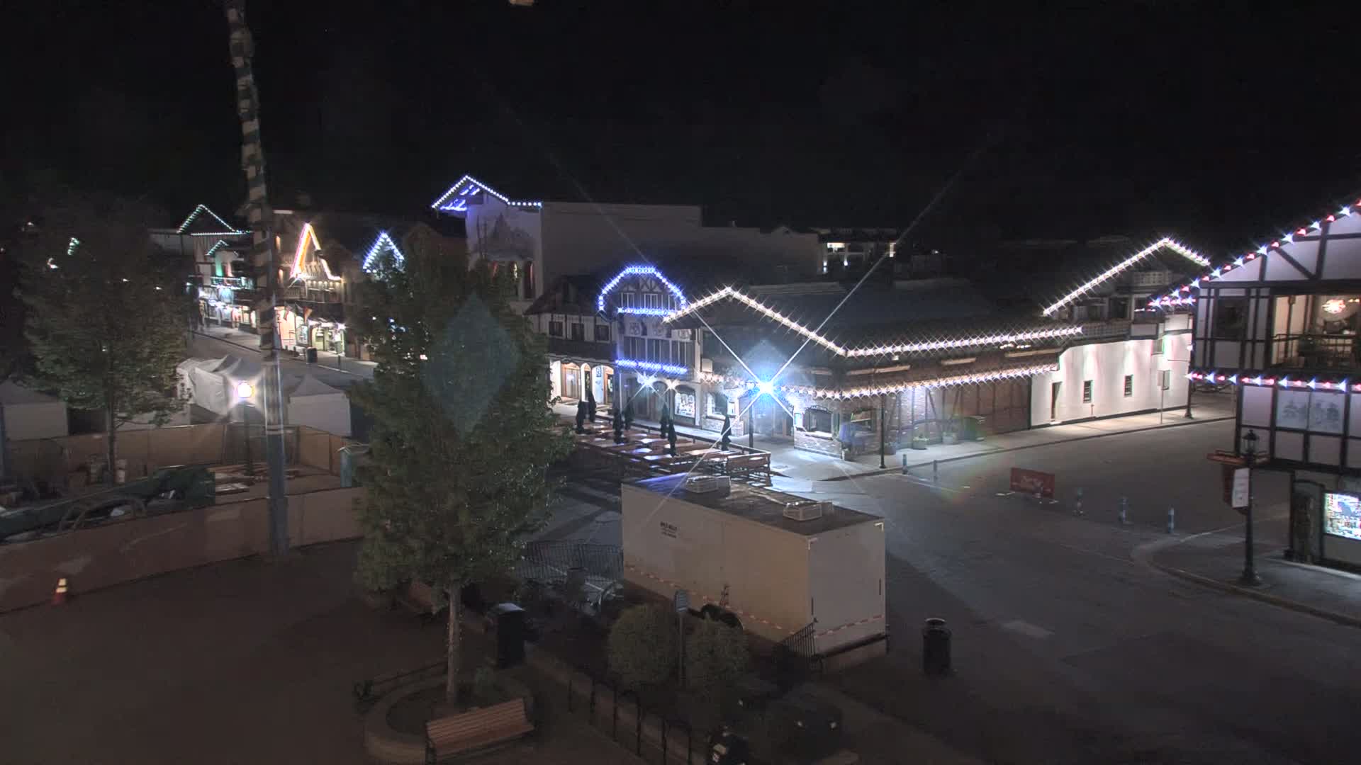 A nighttime view of a street lined with illuminated buildings, some with outdoor seating areas, under a clear, dark sky.