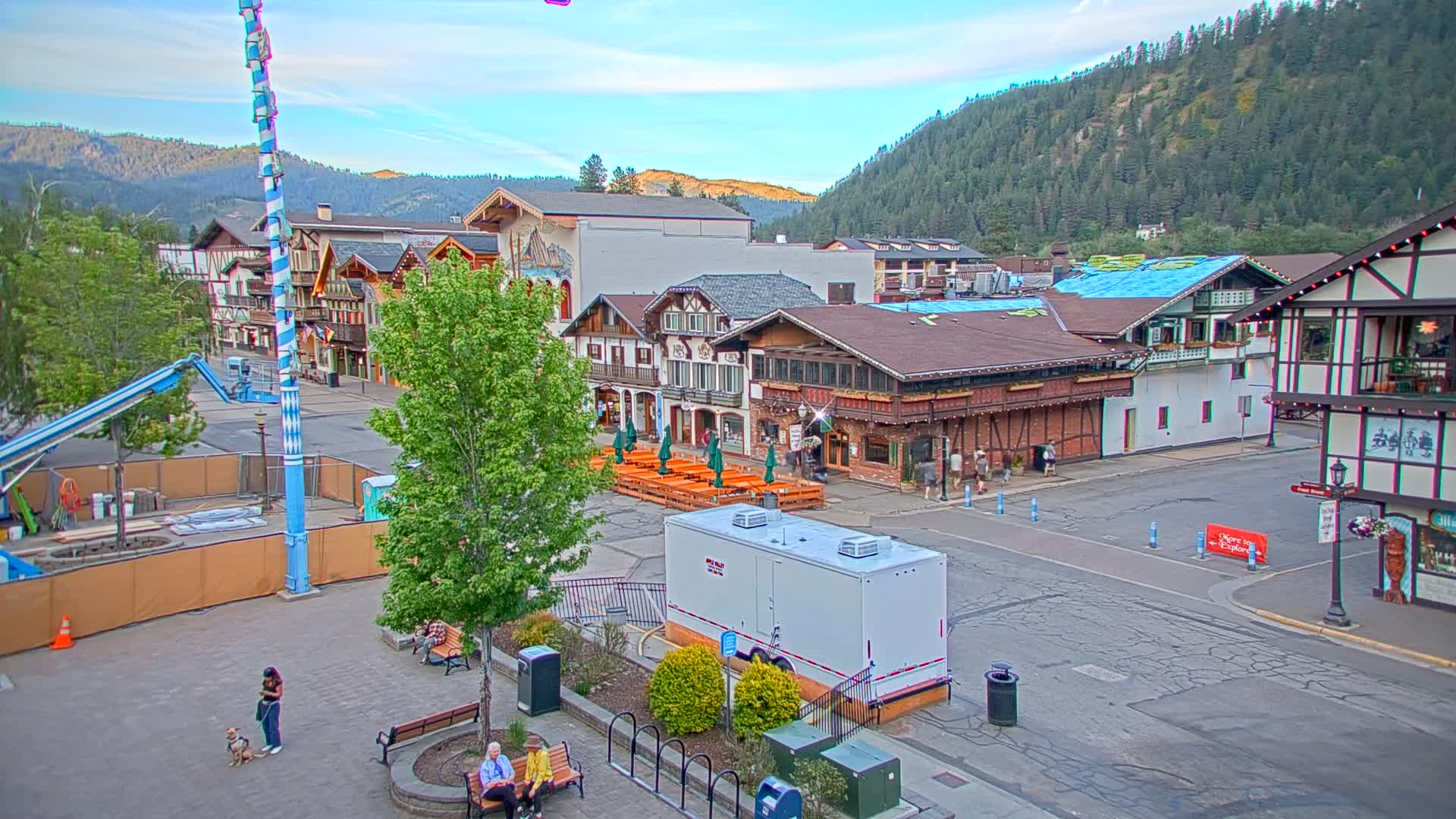 A town square with Bavarian-style buildings, outdoor seating, and a tall maypole, nestled in a valley surrounded by a lush green mountainside under a mostly clear sky.