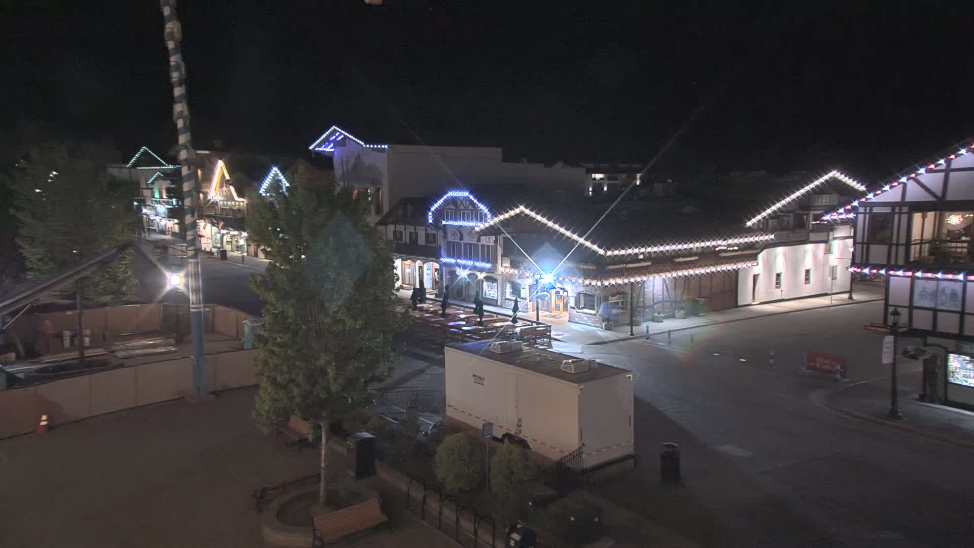 A nighttime view of a street lined with illuminated buildings, a few people are visible, and some construction is underway.