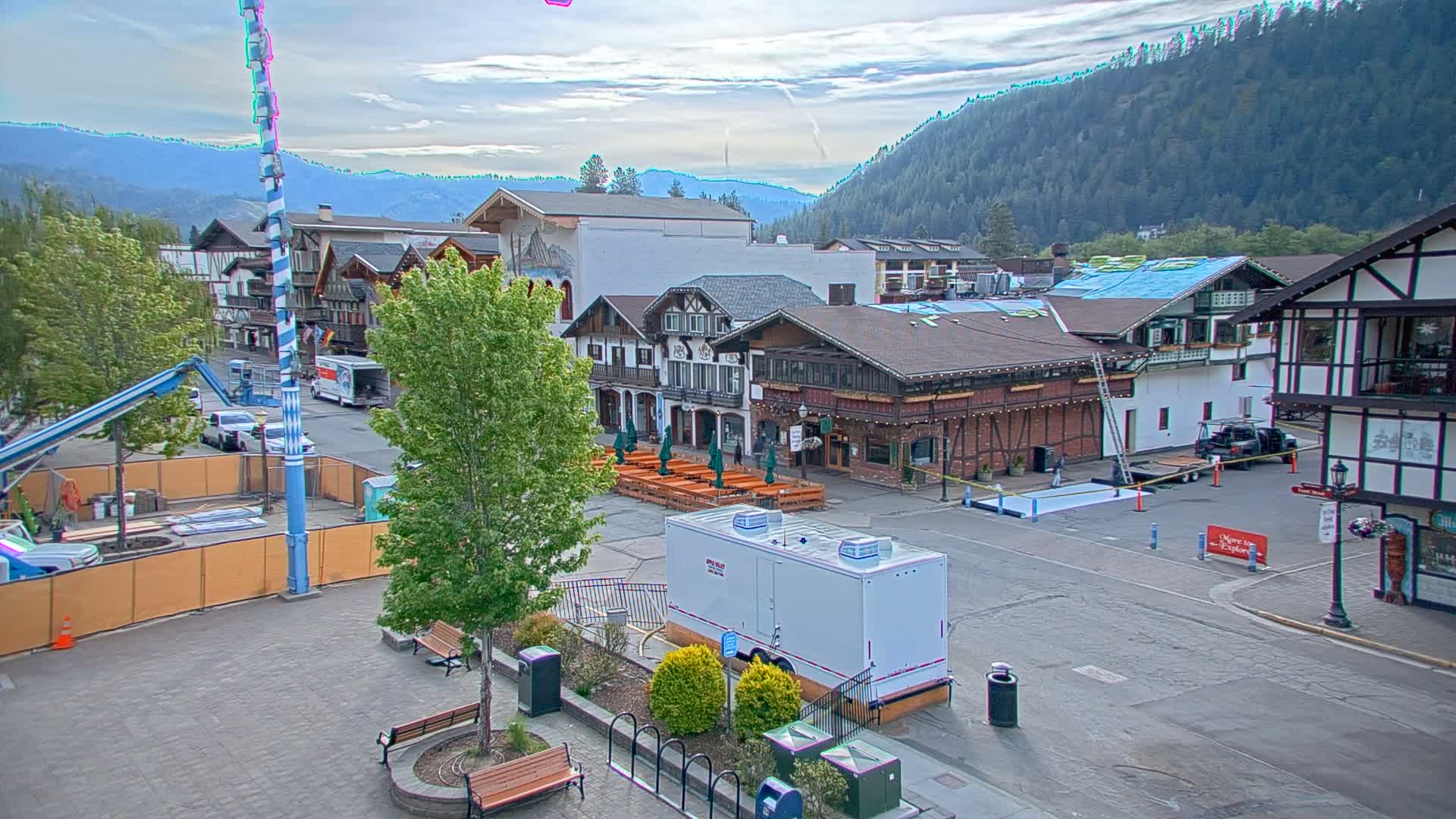 A town square with Bavarian-style buildings, under a partly cloudy sky, is undergoing construction with temporary fencing and equipment visible.