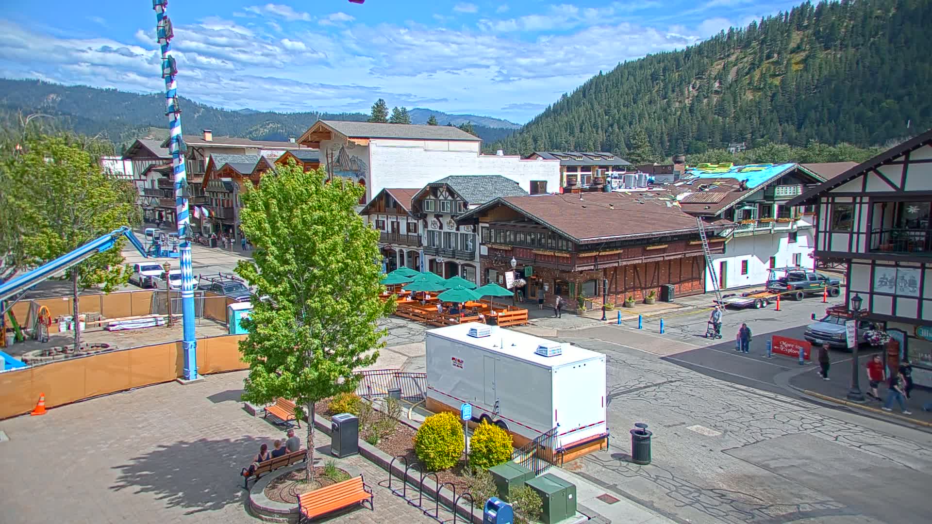 A town square with Bavarian-style buildings, outdoor seating, and construction underway, is situated against a backdrop of a sunny, forested mountainside.
