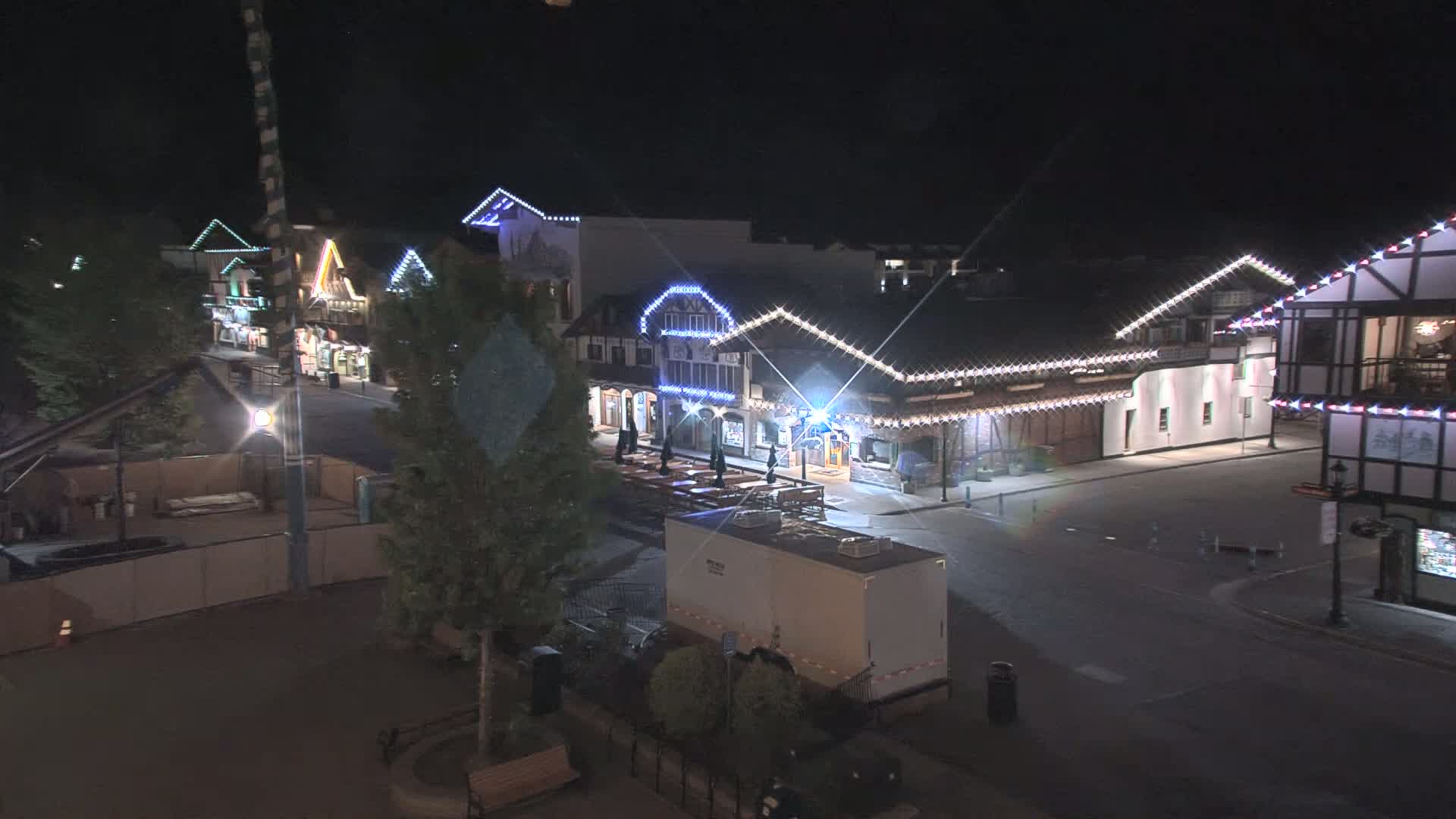 A nighttime view of a street lined with illuminated buildings, featuring outdoor seating and a large trailer parked nearby.