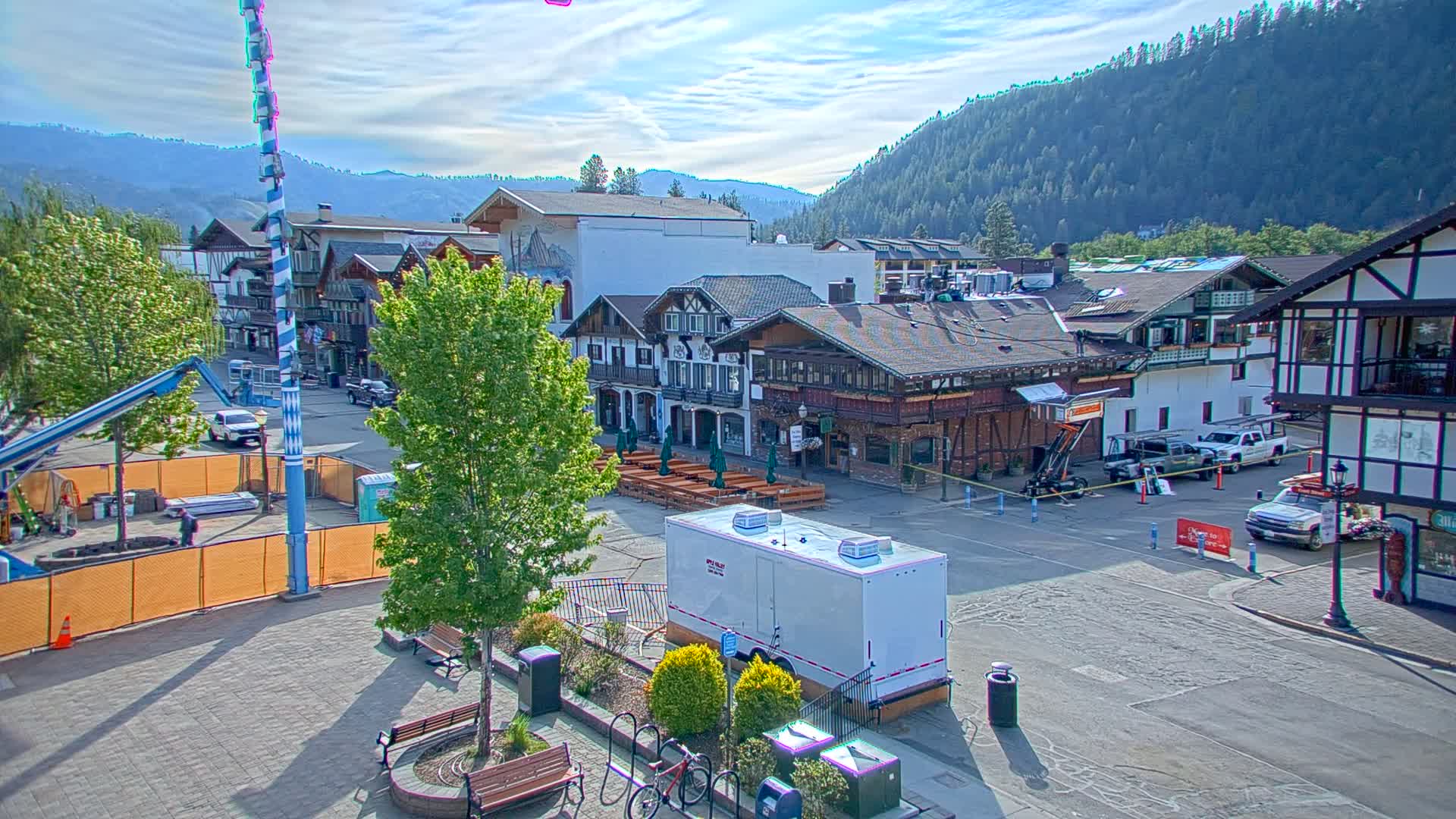 A town square with Bavarian-style buildings, construction equipment, and trees under a mostly sunny sky, surrounded by mountains.