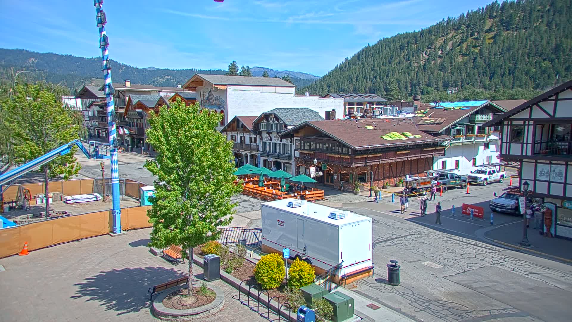 A town square with Bavarian-style buildings, outdoor seating, and a construction site under a sunny sky.