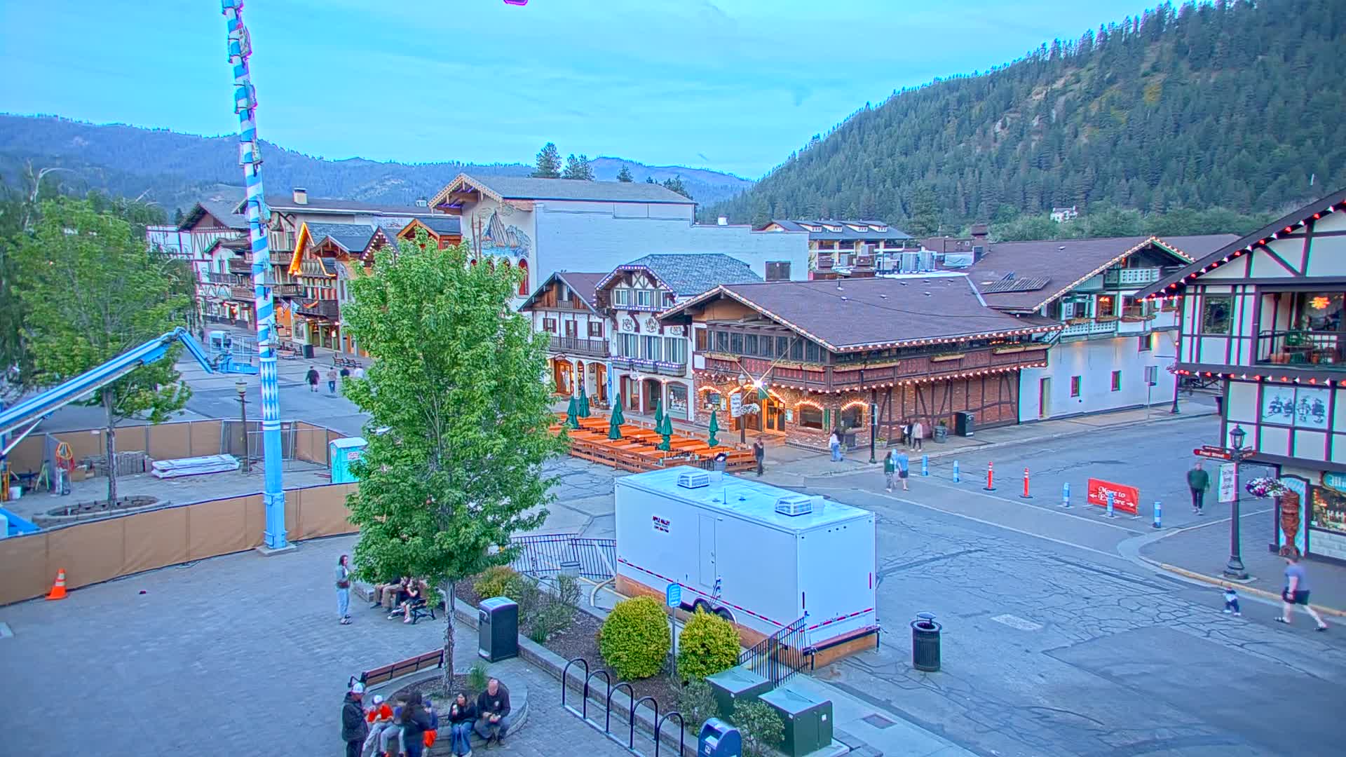 A town square with Bavarian-style buildings, outdoor seating, and a few pedestrians under a twilight sky.