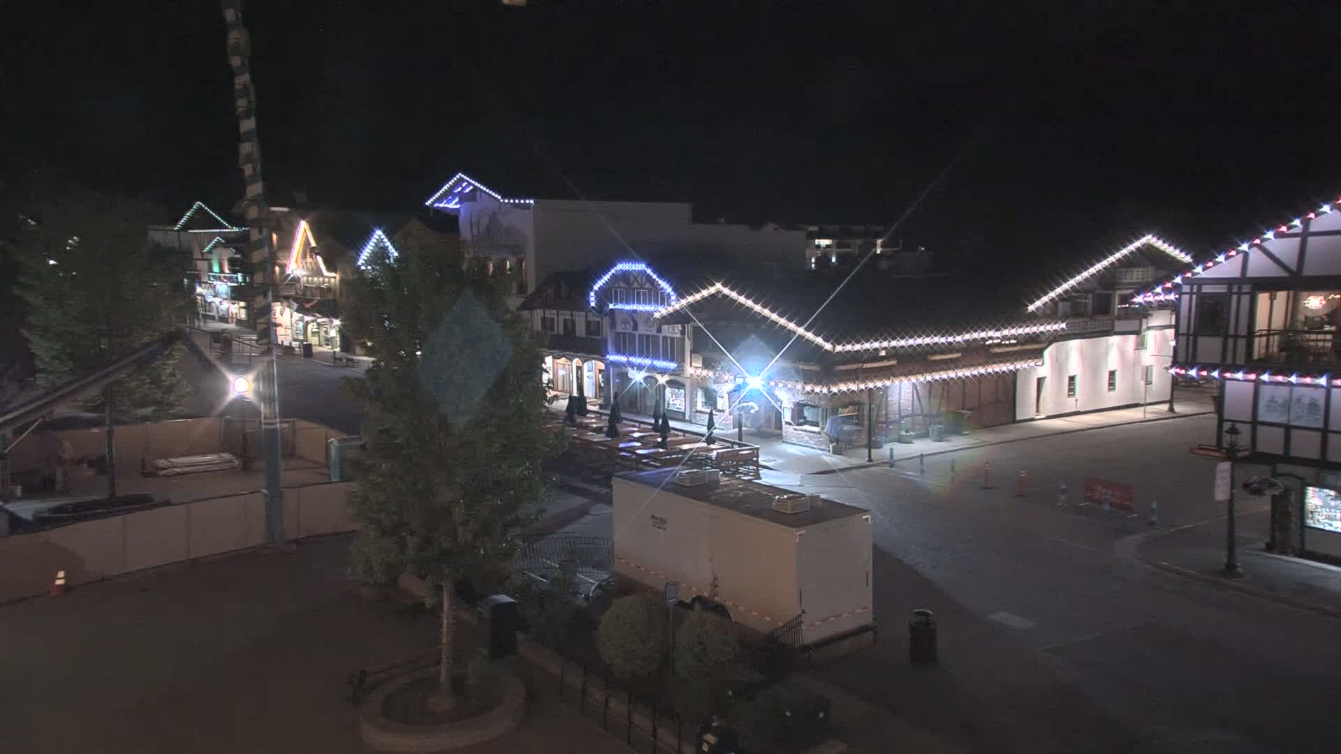A nighttime, mostly empty street lined with illuminated buildings, some with outdoor seating areas, under a clear, dark sky.
