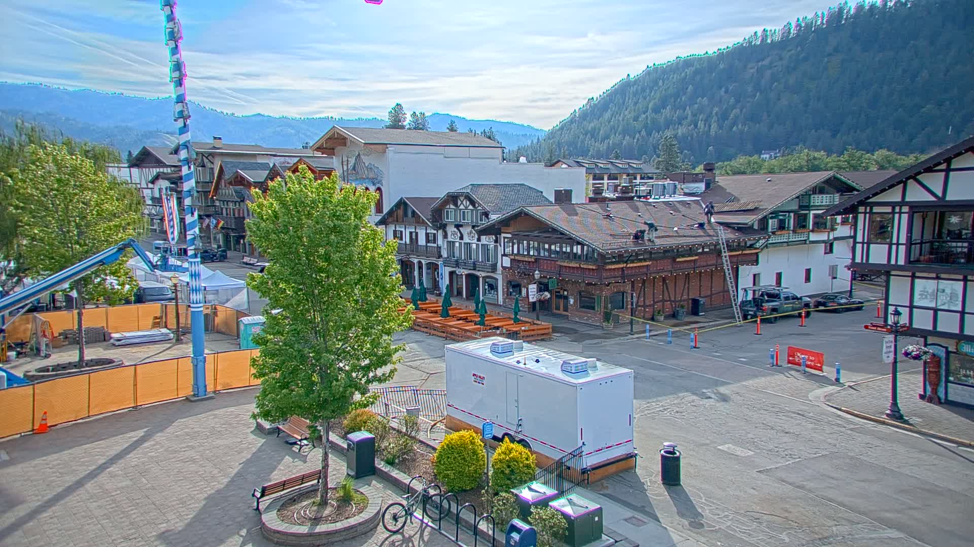 A town square with Bavarian-style buildings, under a mostly sunny sky, is partially closed off for what appears to be a festival or event, with workers on a building's roof.