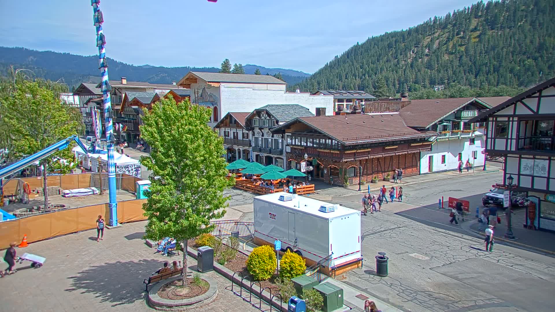 A town square with Bavarian-style buildings, a maypole, and outdoor seating under umbrellas, is set against a backdrop of forested hills under a sunny sky.
