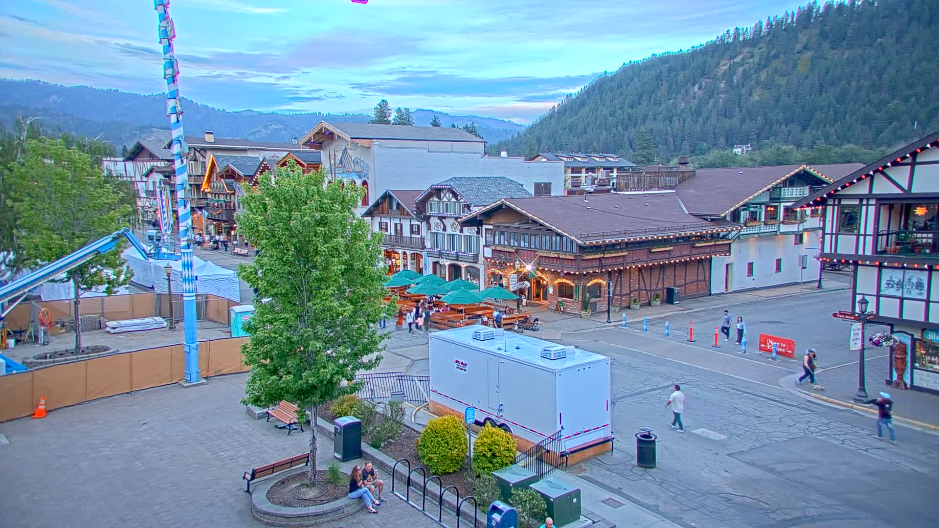 A Bavarian-style village square under a partly cloudy evening sky, with people walking around, outdoor seating at restaurants, and a portable restroom.
