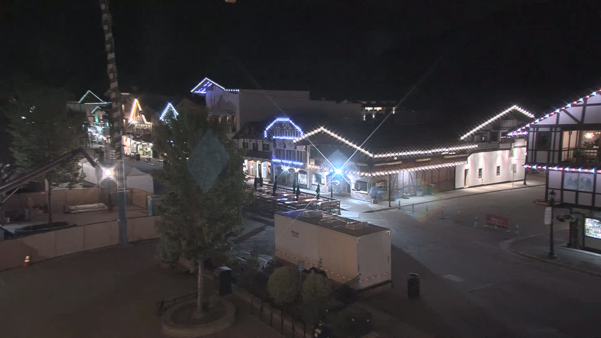 A nighttime view of a street lined with festively lit buildings, a few people are visible, and there is a large trailer in the foreground.