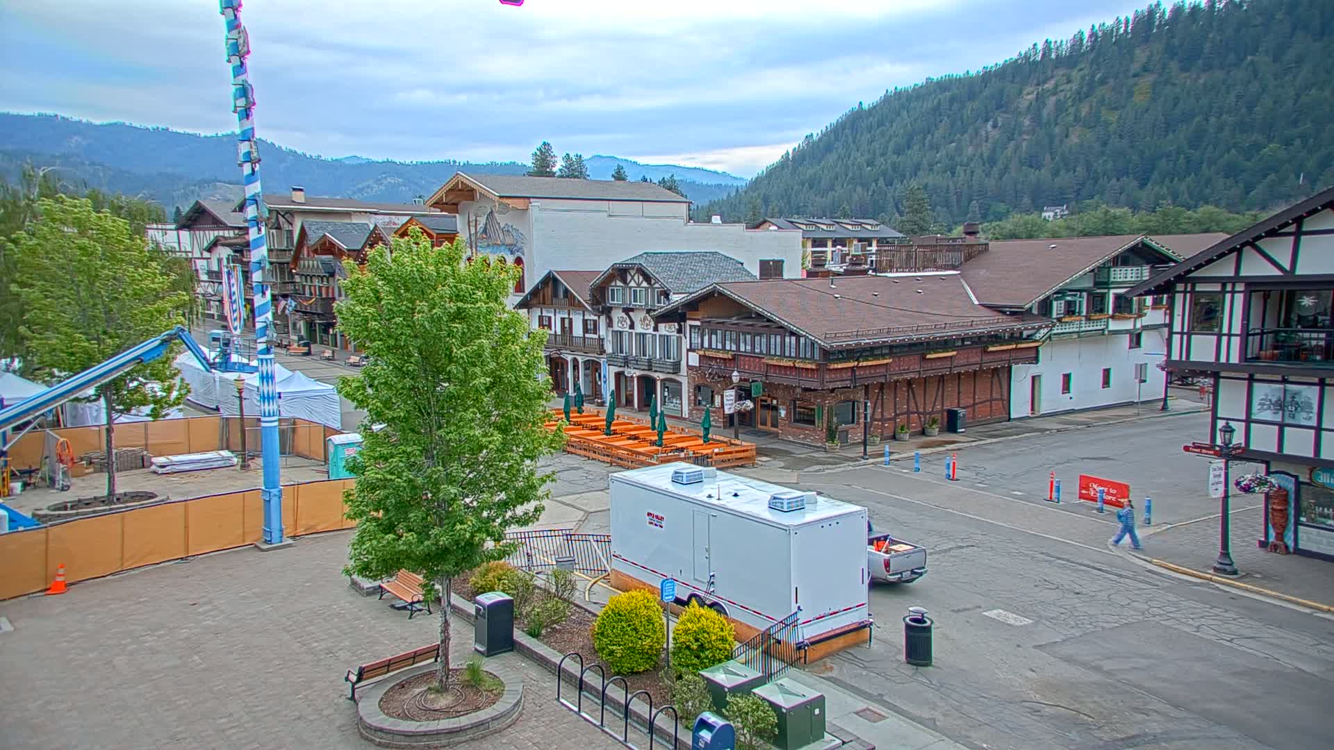 A town square with Bavarian-style buildings, outdoor seating areas, and a tall maypole, under cloudy skies, with a forested mountain in the background.