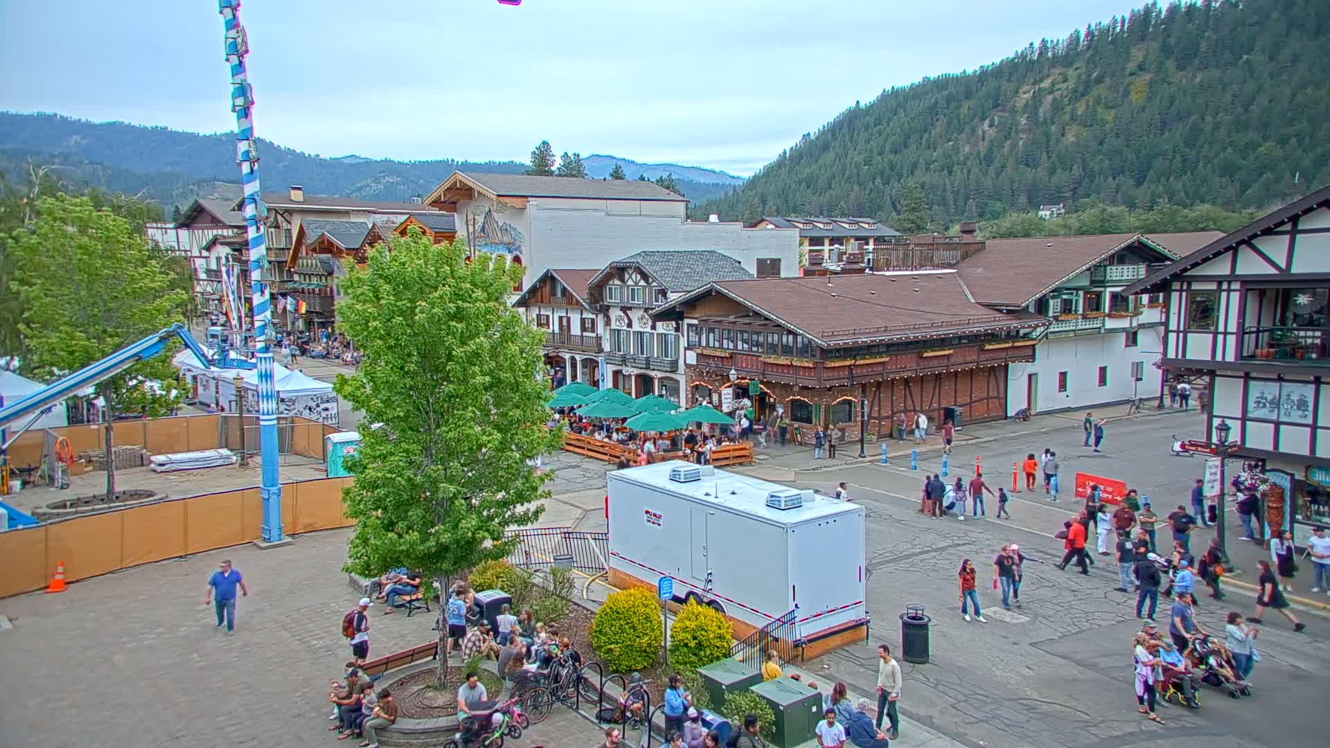 A town square filled with people, surrounded by Bavarian-style buildings, under a partly cloudy sky, with mountains in the background.
