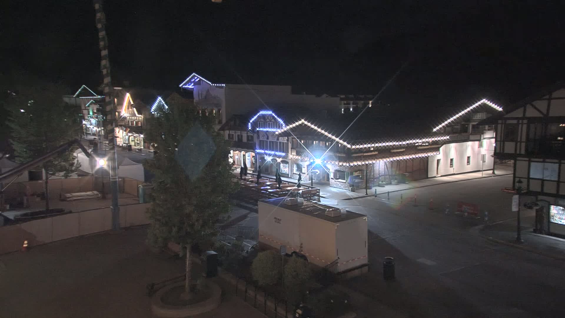 A nighttime street scene shows several buildings decorated with string lights, a few people standing outside, and a large trailer parked on the street.