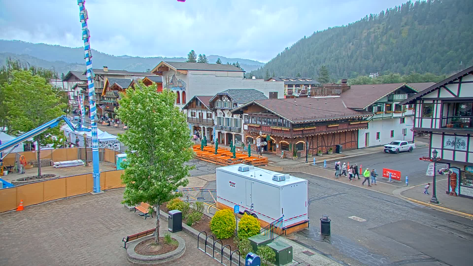 A Bavarian-style town square with outdoor seating, a tall maypole, and construction equipment is shown under an overcast sky, with mountains visible in the background.