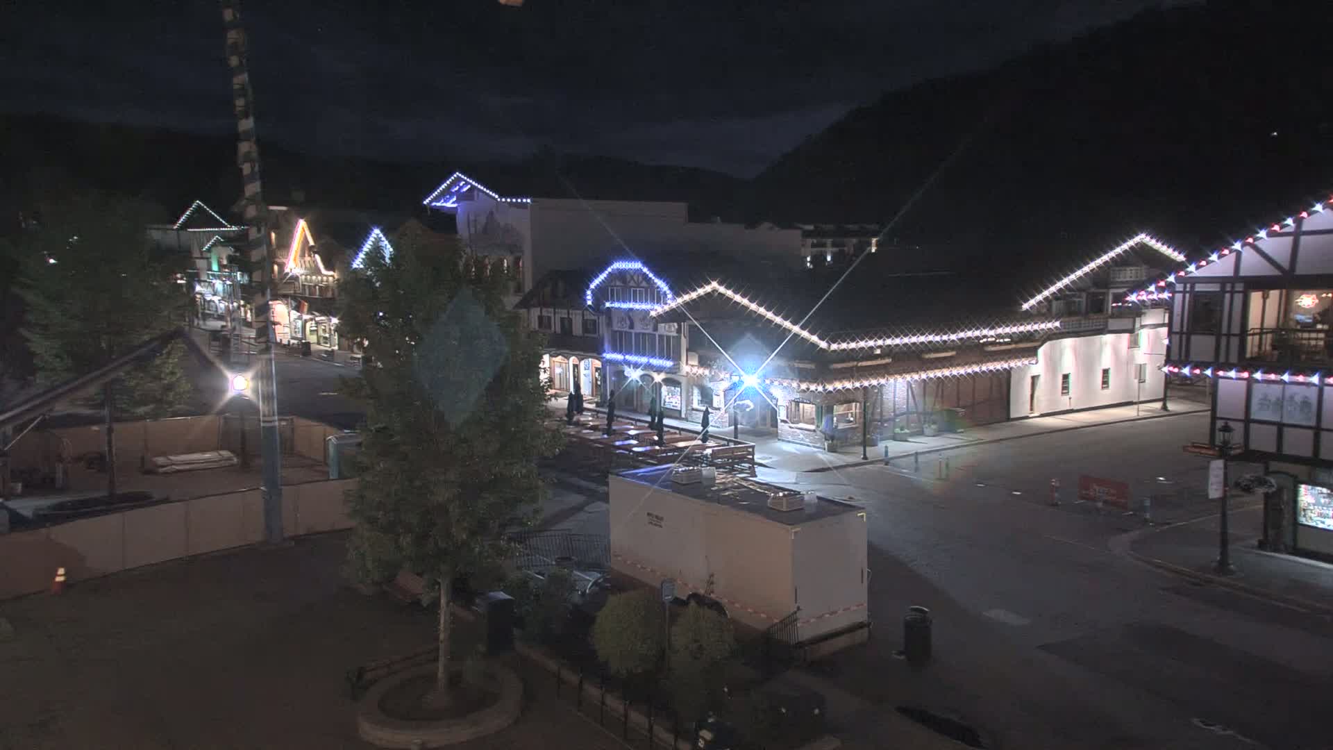 A nighttime view of a town square with several buildings illuminated by string lights, a few trees, and a large, empty parking lot.