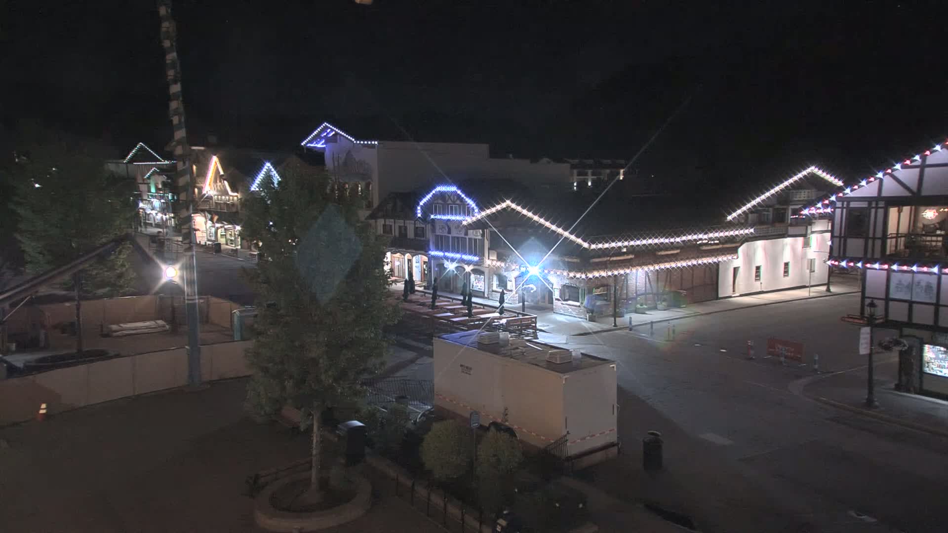 A nighttime view of a mostly empty street lined with festively lit buildings, a large trailer is parked nearby.