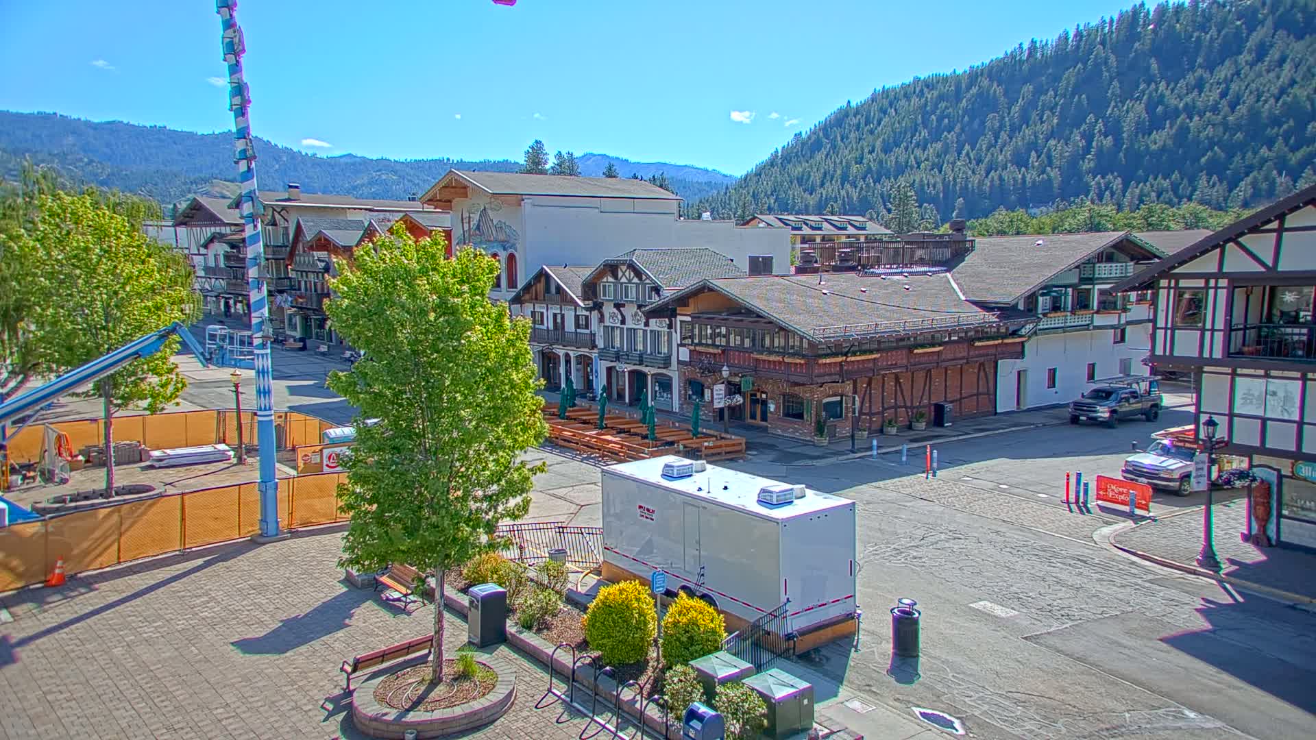 A town square with several buildings in a Bavarian architectural style sits against a backdrop of forested mountains under a clear, sunny sky.