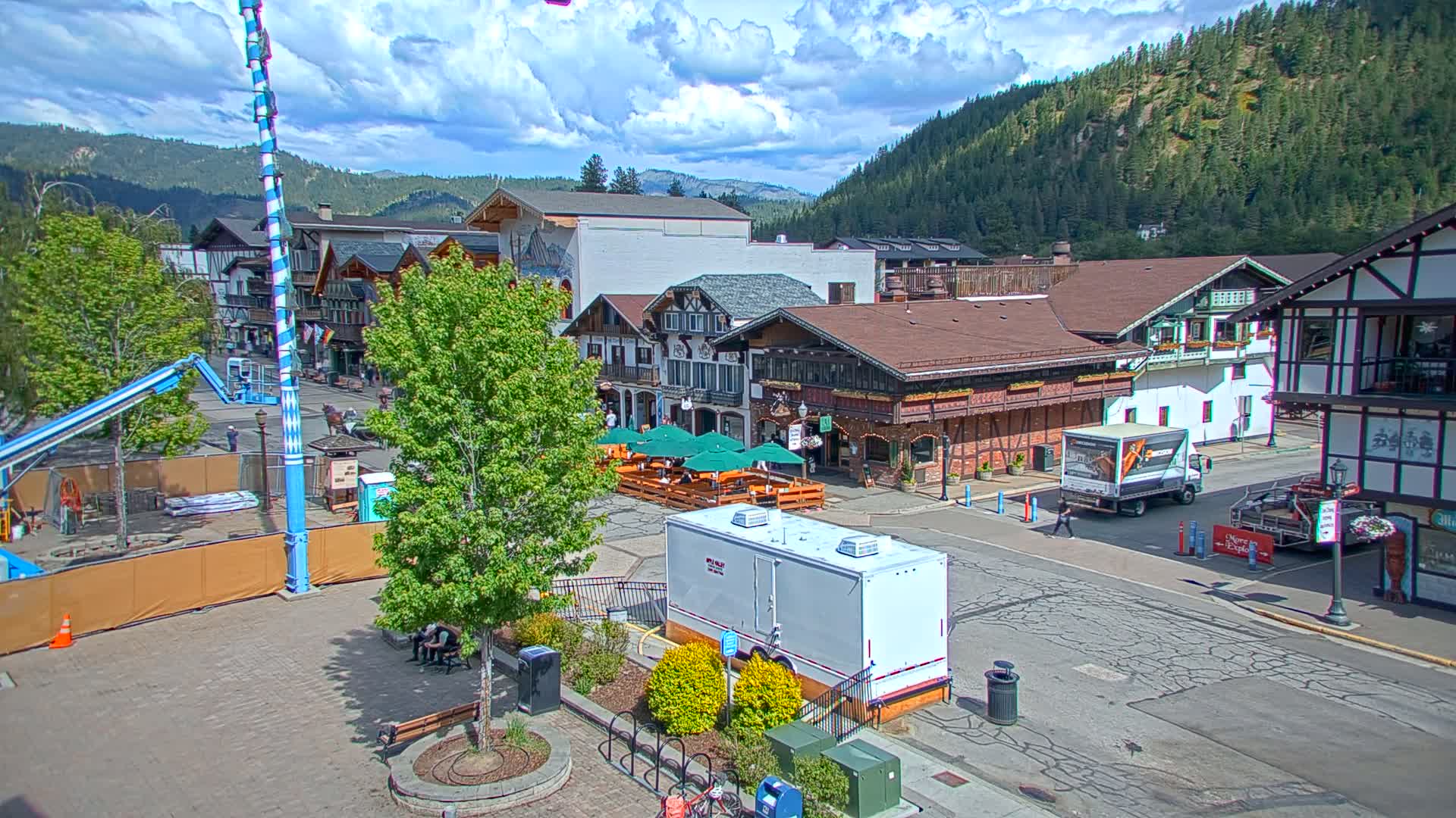 A partly cloudy day reveals a Bavarian-style village street scene nestled against a mountain backdrop, with outdoor seating areas at restaurants and a mobile restroom visible in the square.