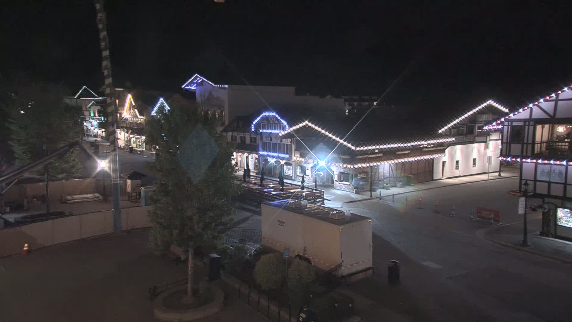 A nighttime, brightly-lit town square with several buildings decorated with string lights, some outdoor seating, and a large portable restroom.