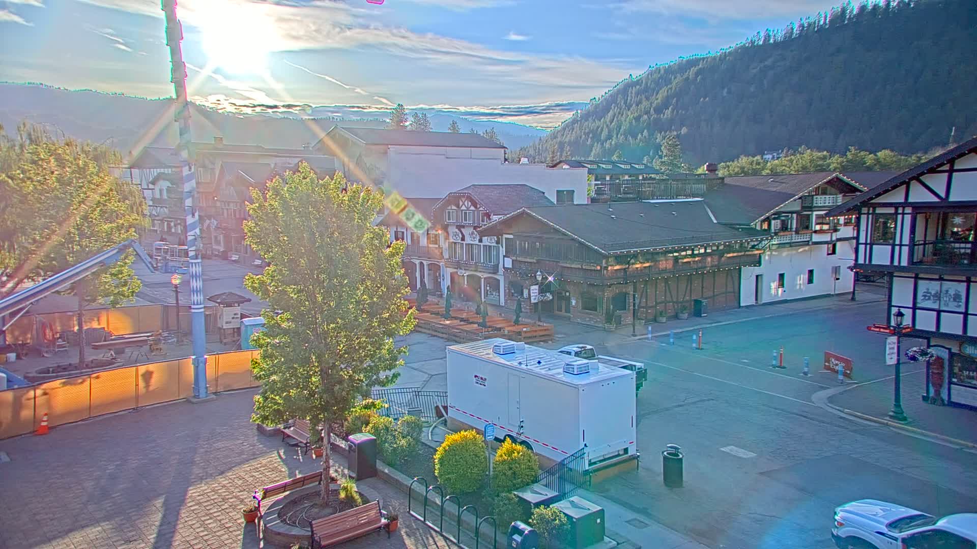 A sunlit town square with several buildings, including some with half-timbered architecture, sits in a valley surrounded by forested hills under a mostly clear sky.