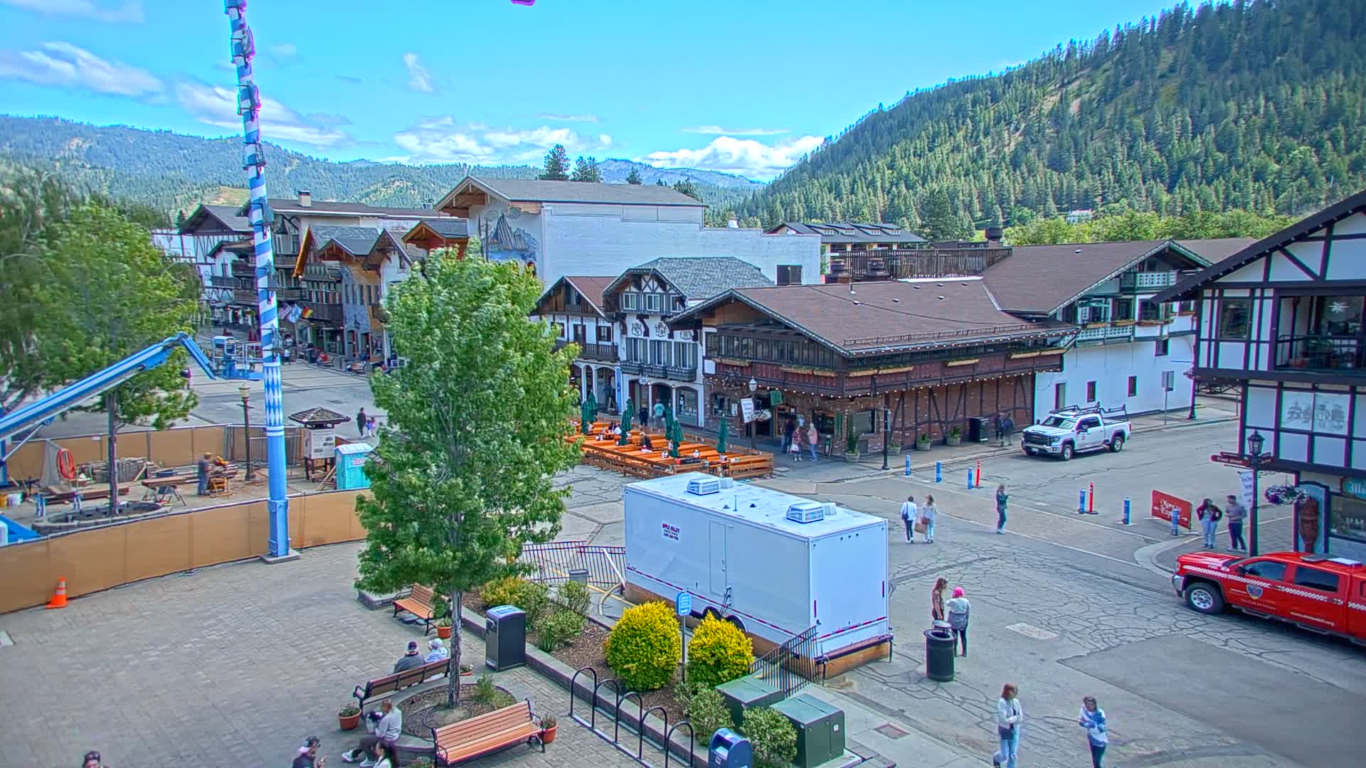 A mostly sunny day reveals a town square with Bavarian-style buildings, outdoor seating areas, and several people walking around, all nestled at the base of a forested mountain range.