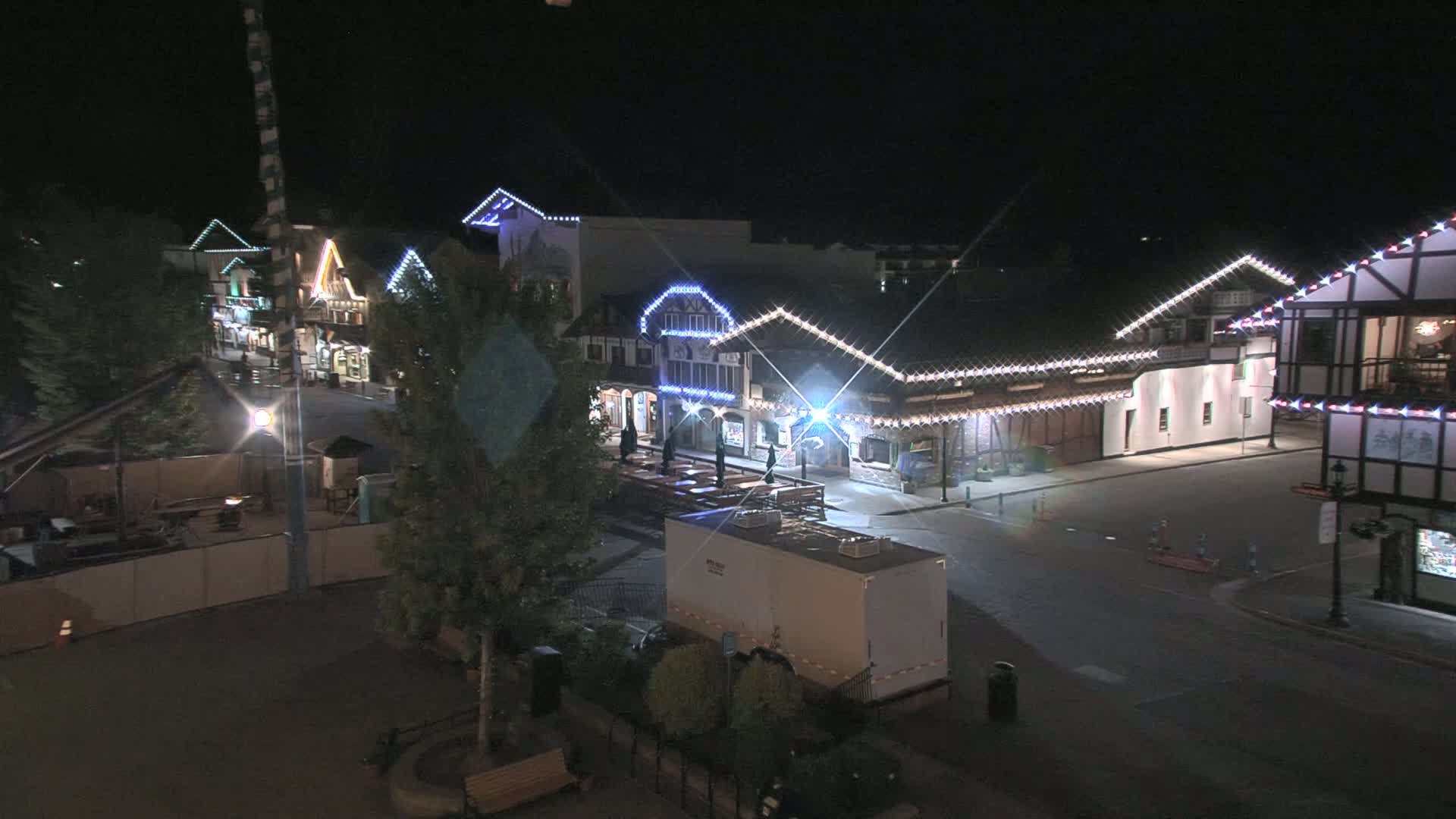 A nighttime, overhead view of a town square with several illuminated buildings, featuring string lights and a few sparsely populated outdoor seating areas.