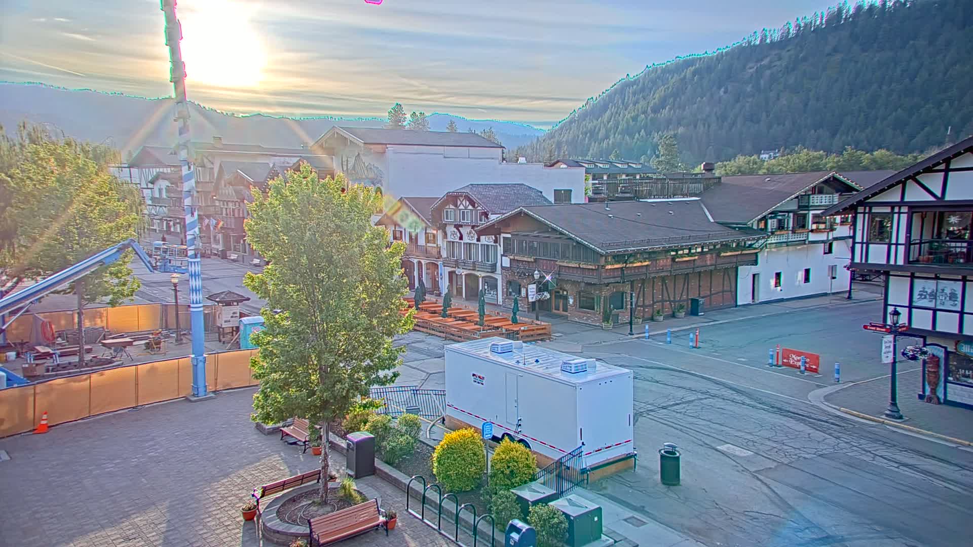A sunlit town square with Bavarian-style buildings, a portable restroom, and a partially constructed fairground or similar event space against a backdrop of forested mountains.