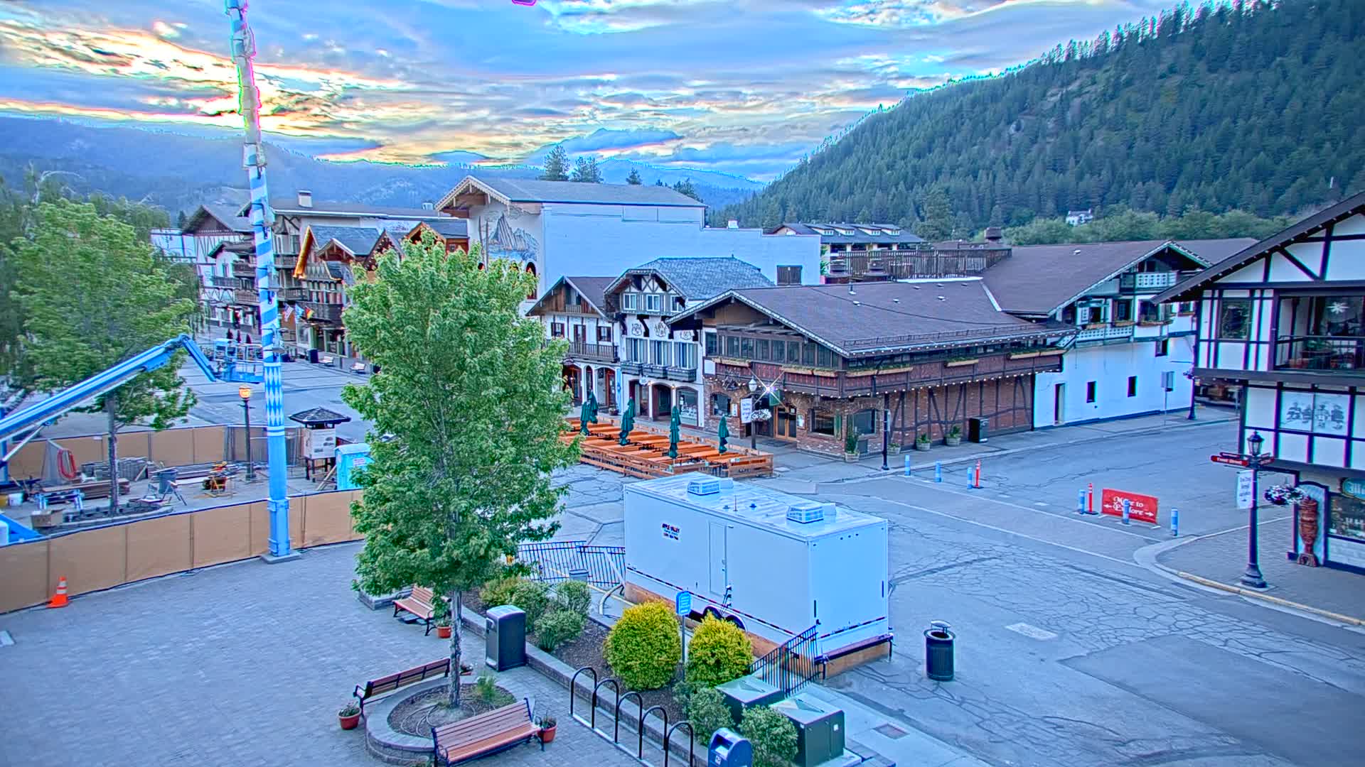 A town square with Bavarian-style buildings and a mountain backdrop under a partly cloudy sunrise sky.