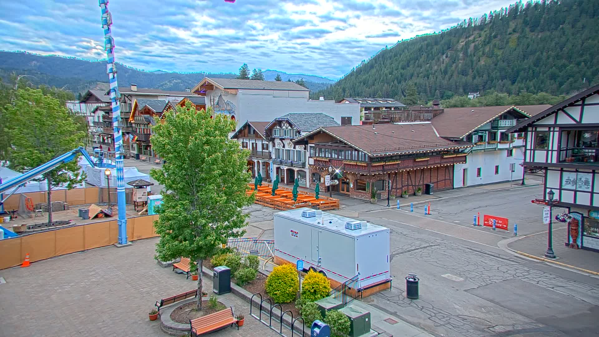 A town square with Bavarian-style buildings, outdoor seating, and construction equipment under a partly cloudy sky, set against a backdrop of forested mountains.
