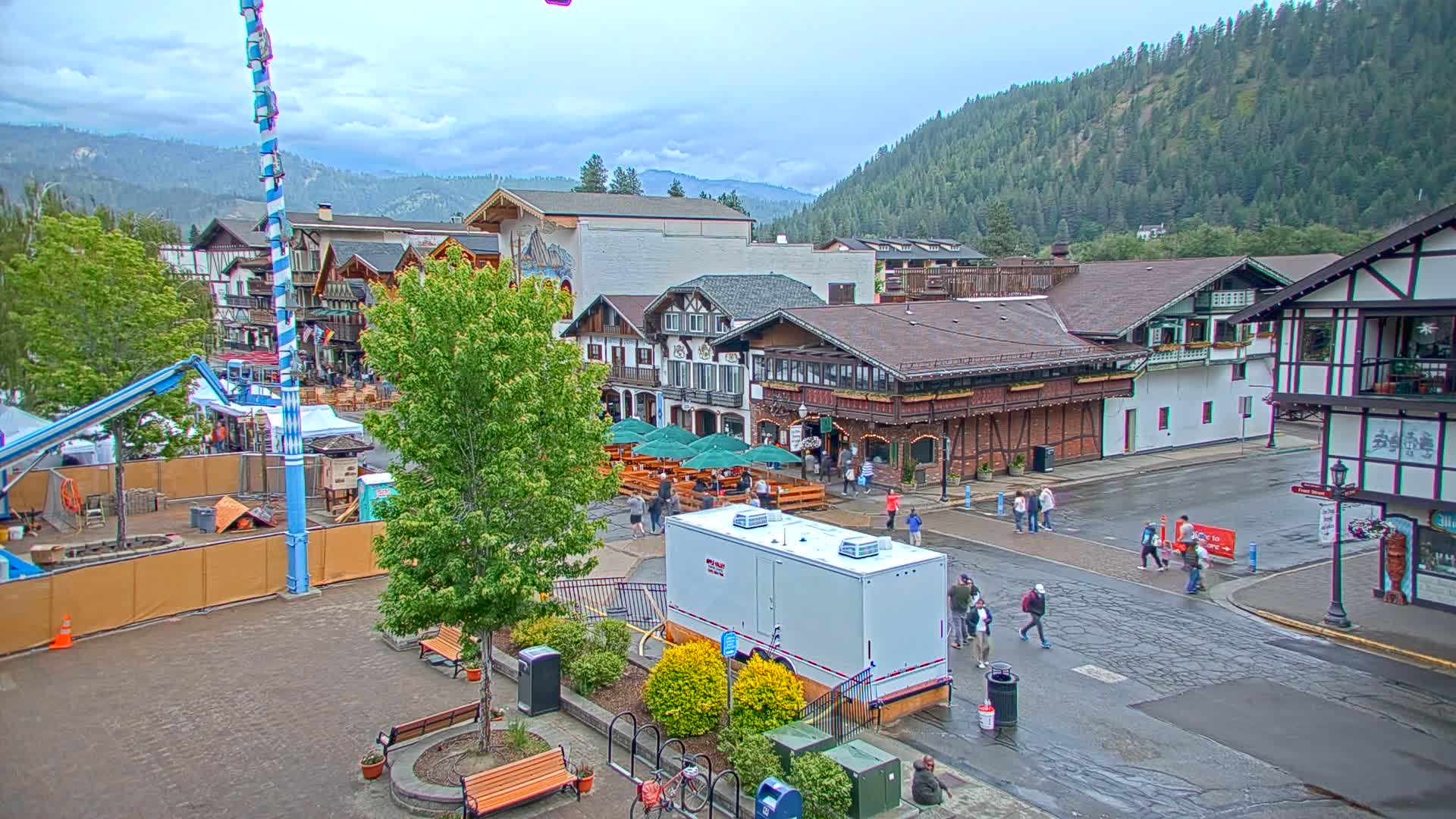 A town square with Bavarian-style buildings, outdoor seating areas, and people walking on a wet street under an overcast sky.