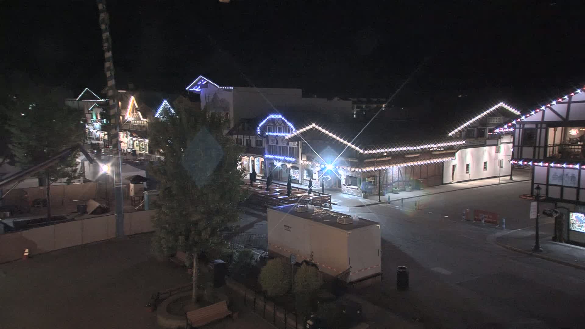 A nighttime, illuminated street scene shows several buildings decorated with string lights, a large empty area, and a few trees.