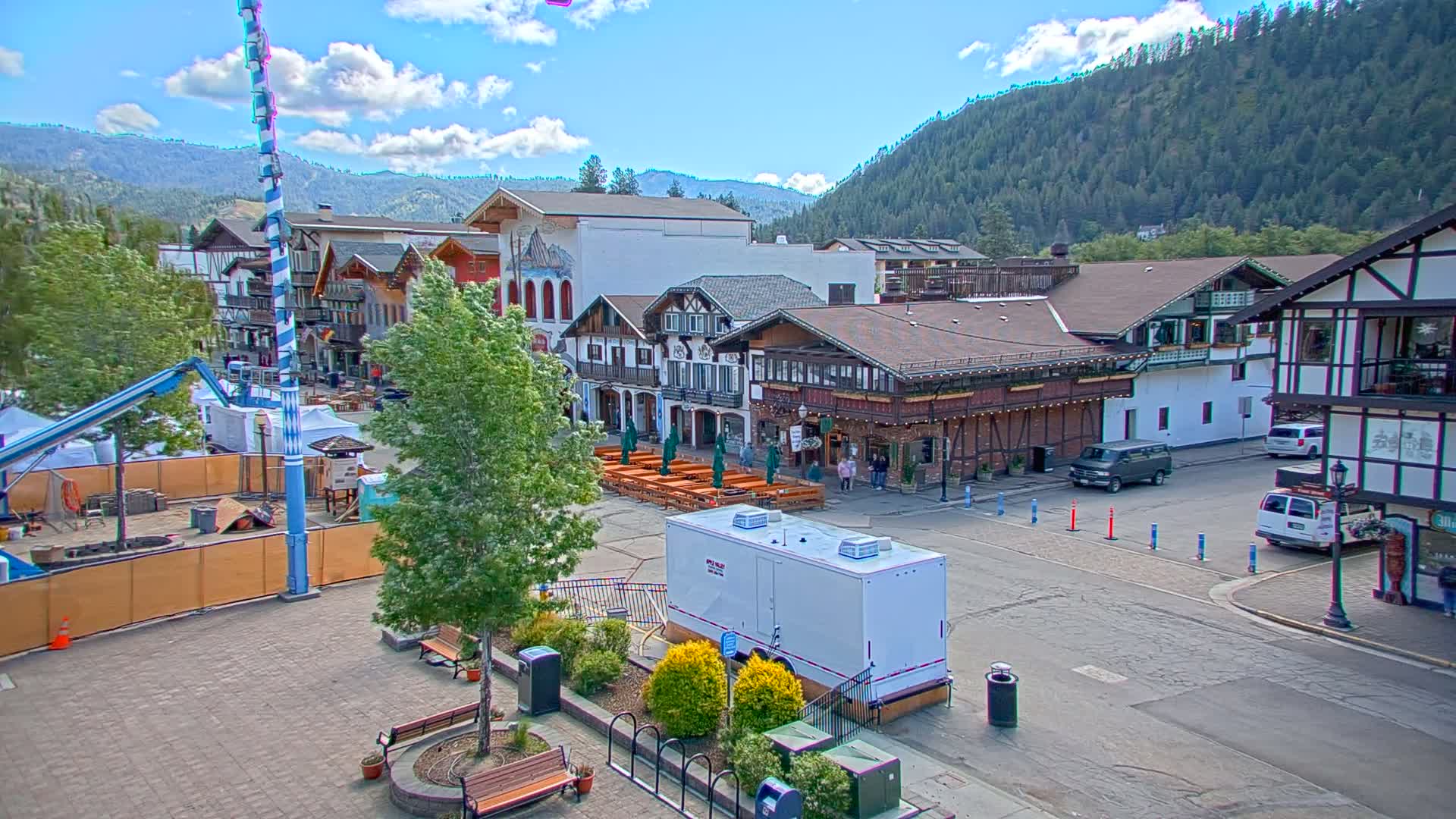A town square with Bavarian-style buildings, outdoor seating areas, and a few vehicles, under a partly cloudy sky with mountains in the background.