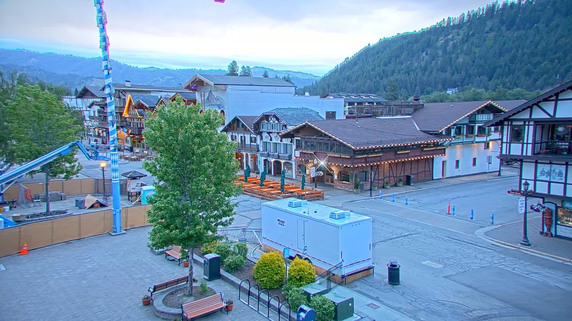 A paved town square with Bavarian-style buildings, outdoor seating, and trees under a cloudy twilight sky, nestled against a forested mountainside.