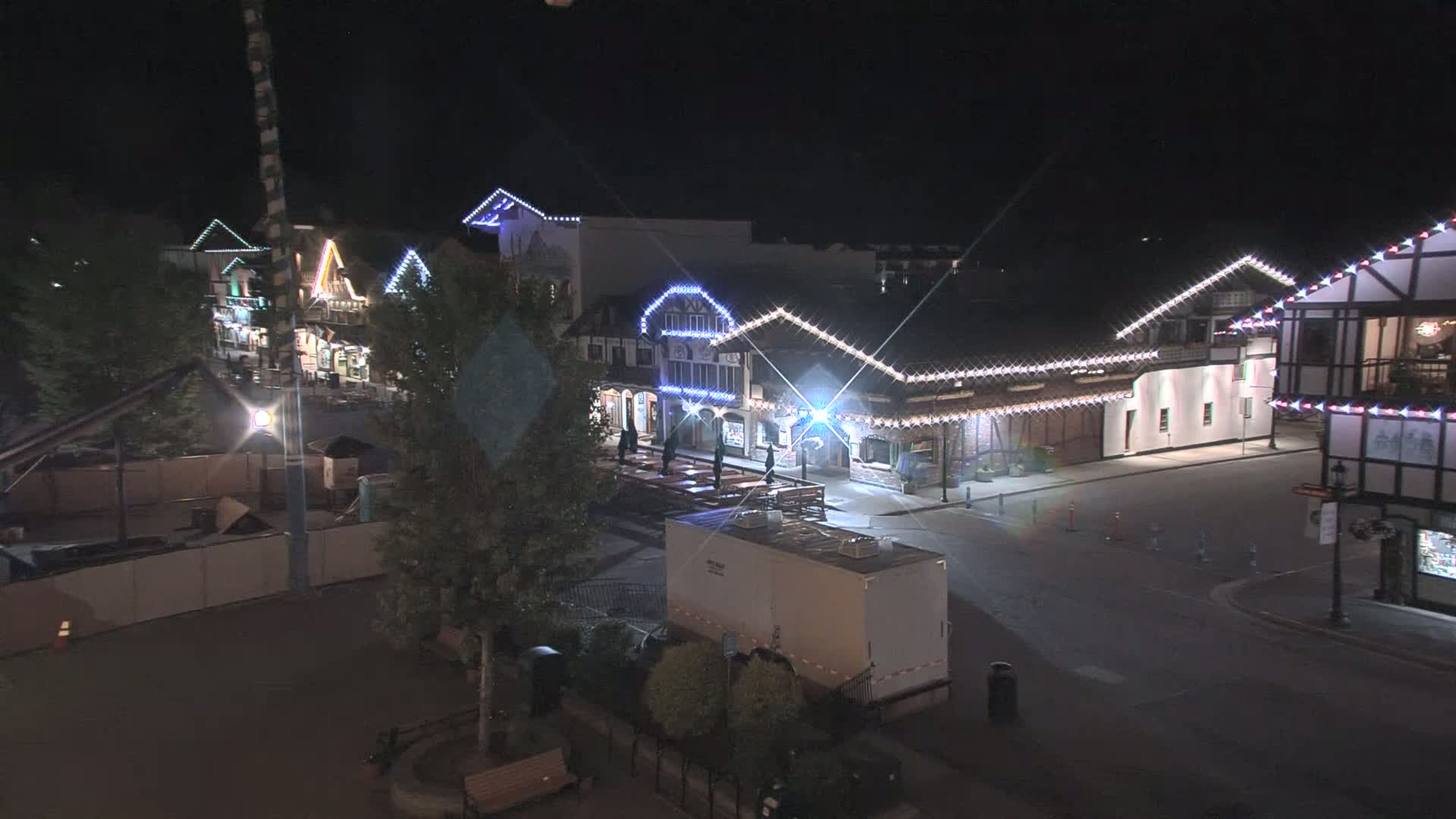 A nighttime, brightly-lit village square features several buildings decorated with string lights, with a few people visible and a portable restroom parked nearby.