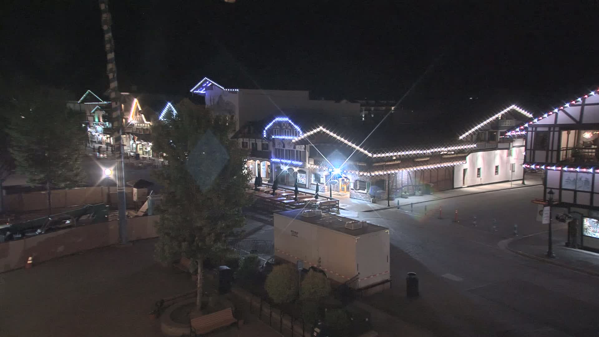 A nighttime, outdoor scene shows a row of illuminated buildings with string lights, a mostly empty street, and some trees.
