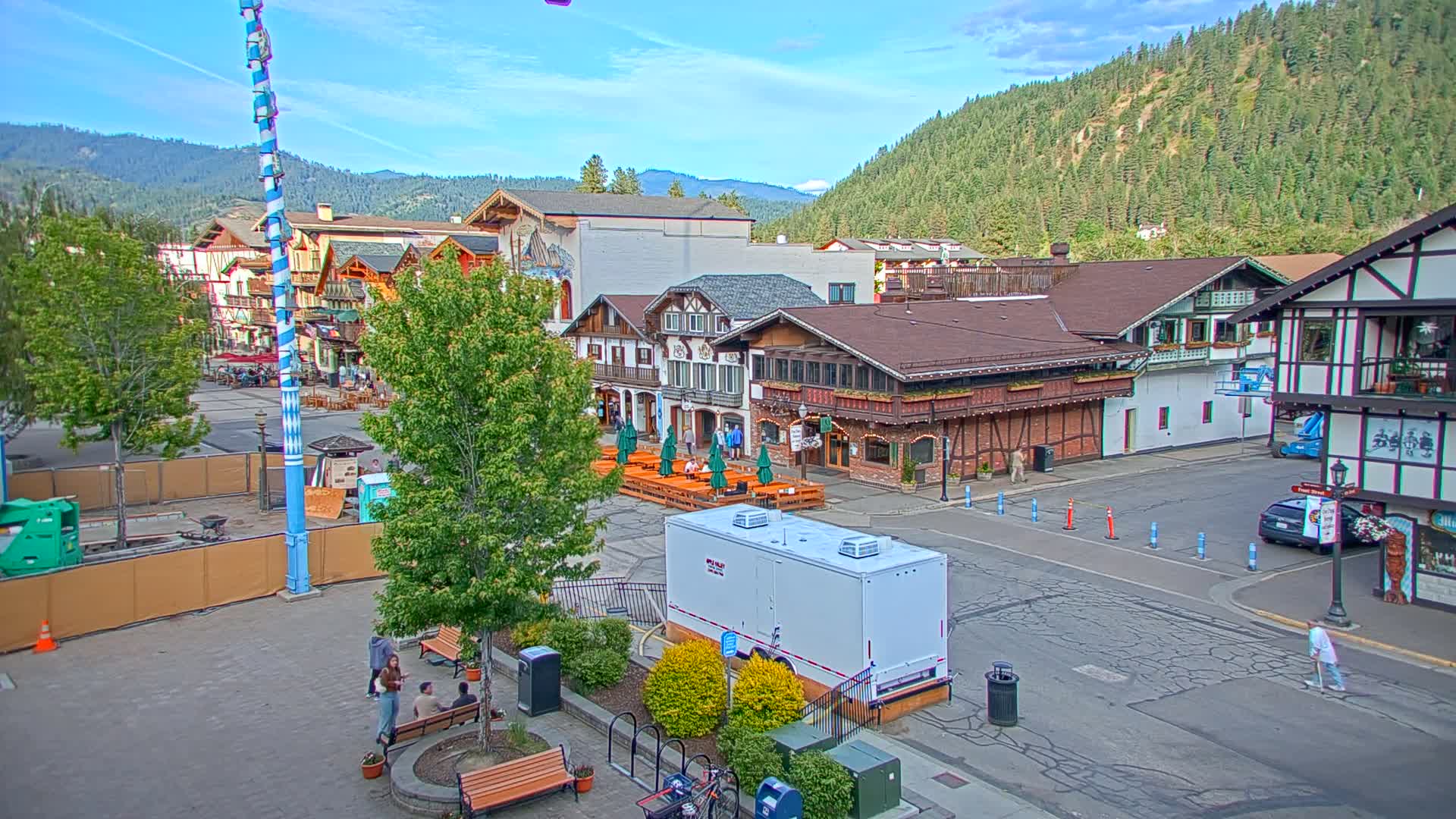 A town square with Bavarian-style buildings, outdoor seating, and a tall maypole is seen on a sunny day with mountains in the background.