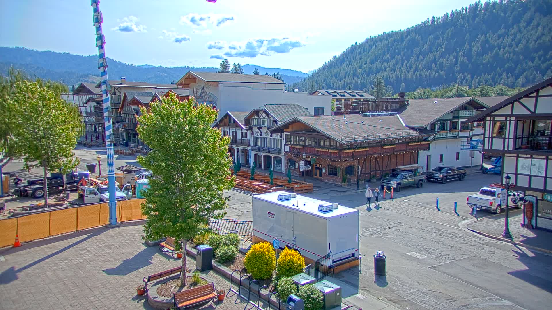 A town square with Bavarian-style buildings, a maypole, and several vehicles under a mostly sunny sky, backed by a mountain range covered in lush green trees.